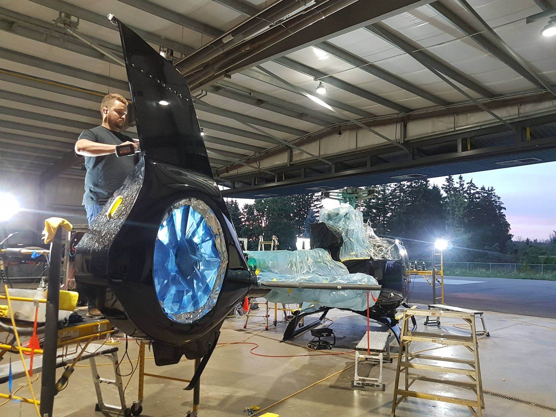 Man working on a large, dark, ornate sculpture with a blue light inside, in a hangar.