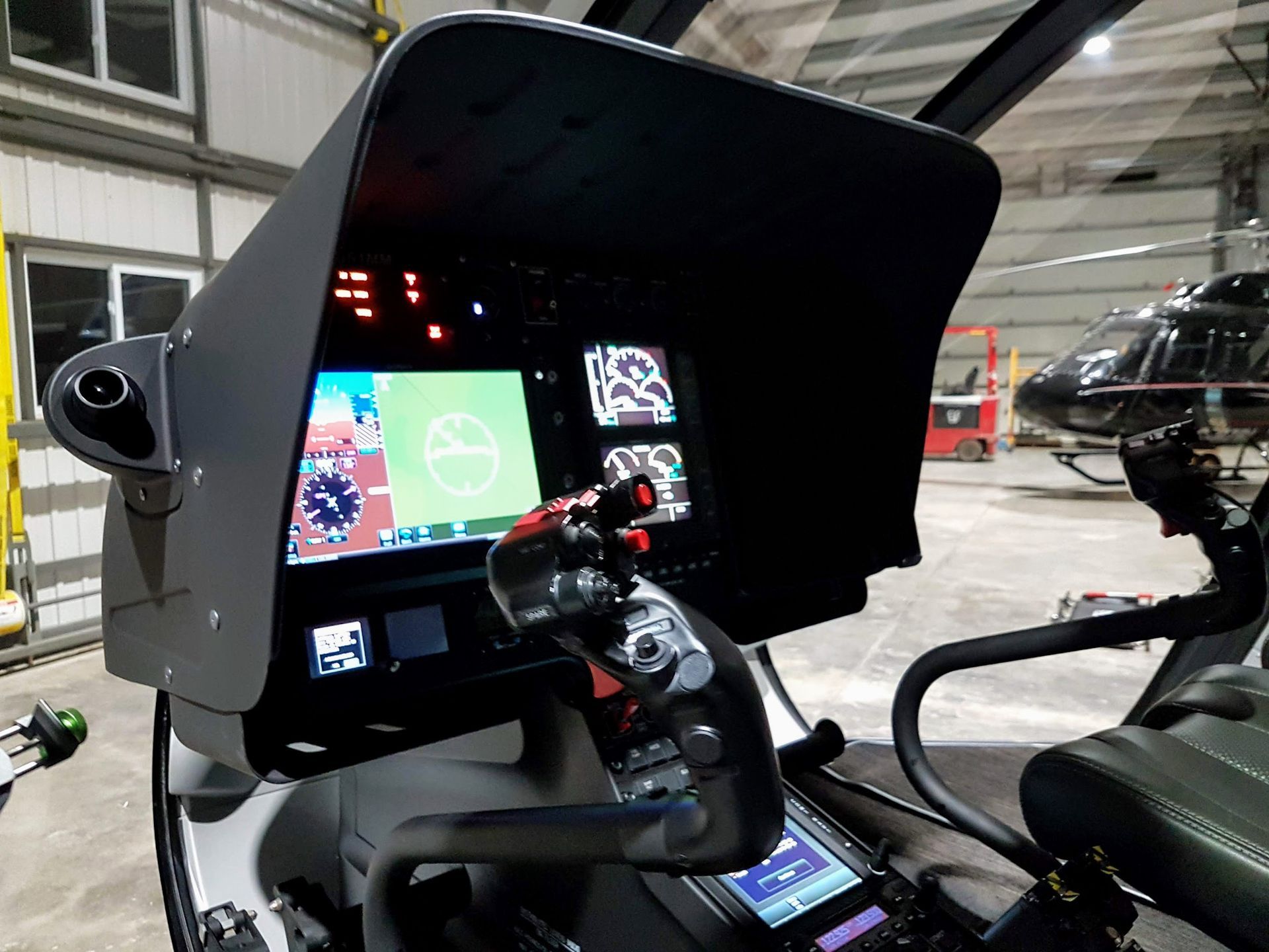 Helicopter cockpit: displays of flight data, control stick, and overhead panels. Black, gray, and red colors.