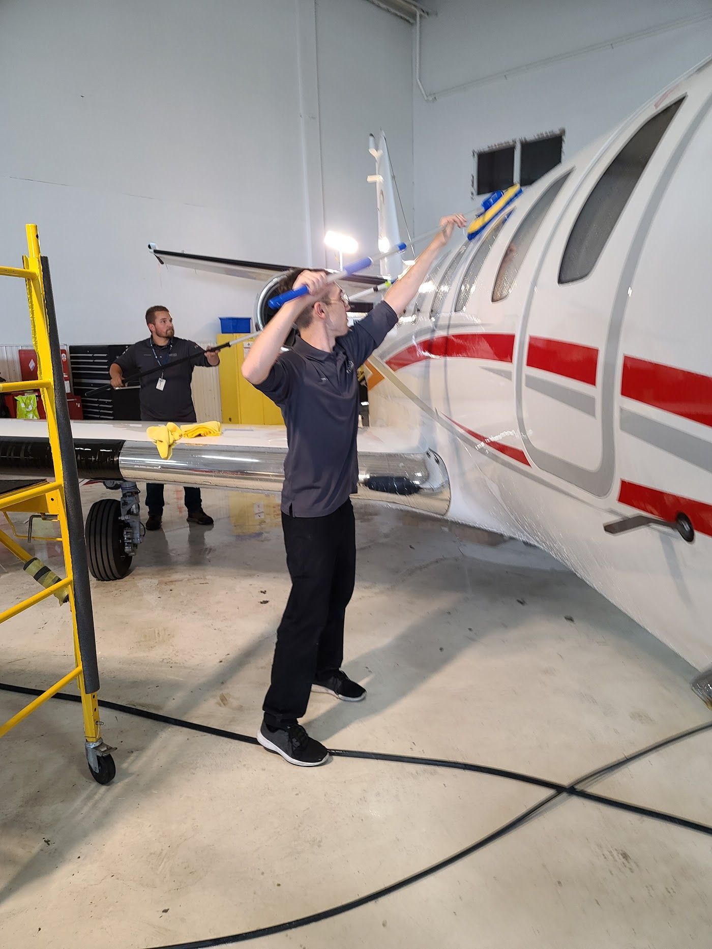 Two people working on a small, white and red airplane in a hangar. One uses a tool on the window.