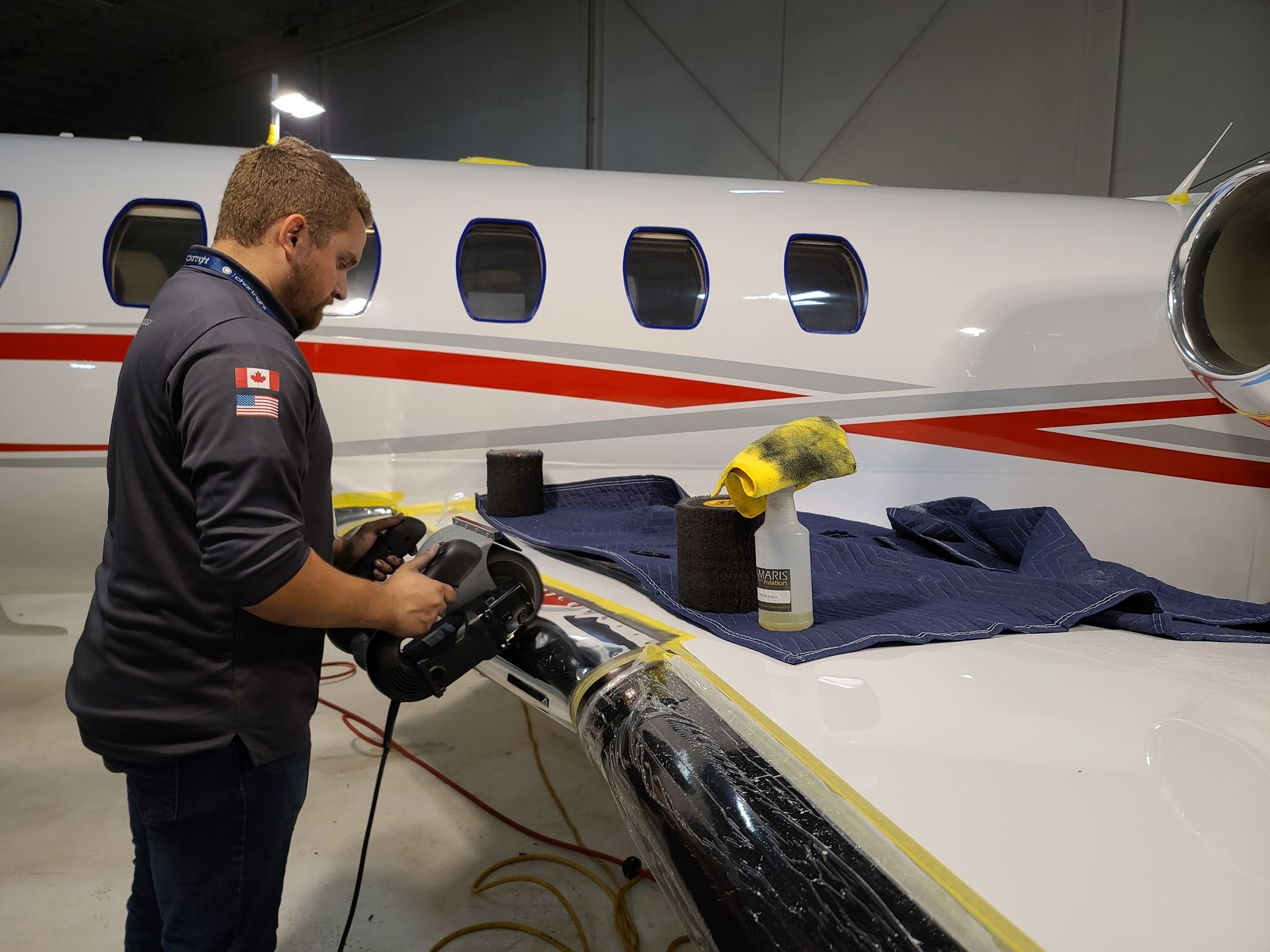 Man using a power tool on an airplane wing in a hangar, wing is masked with tape.