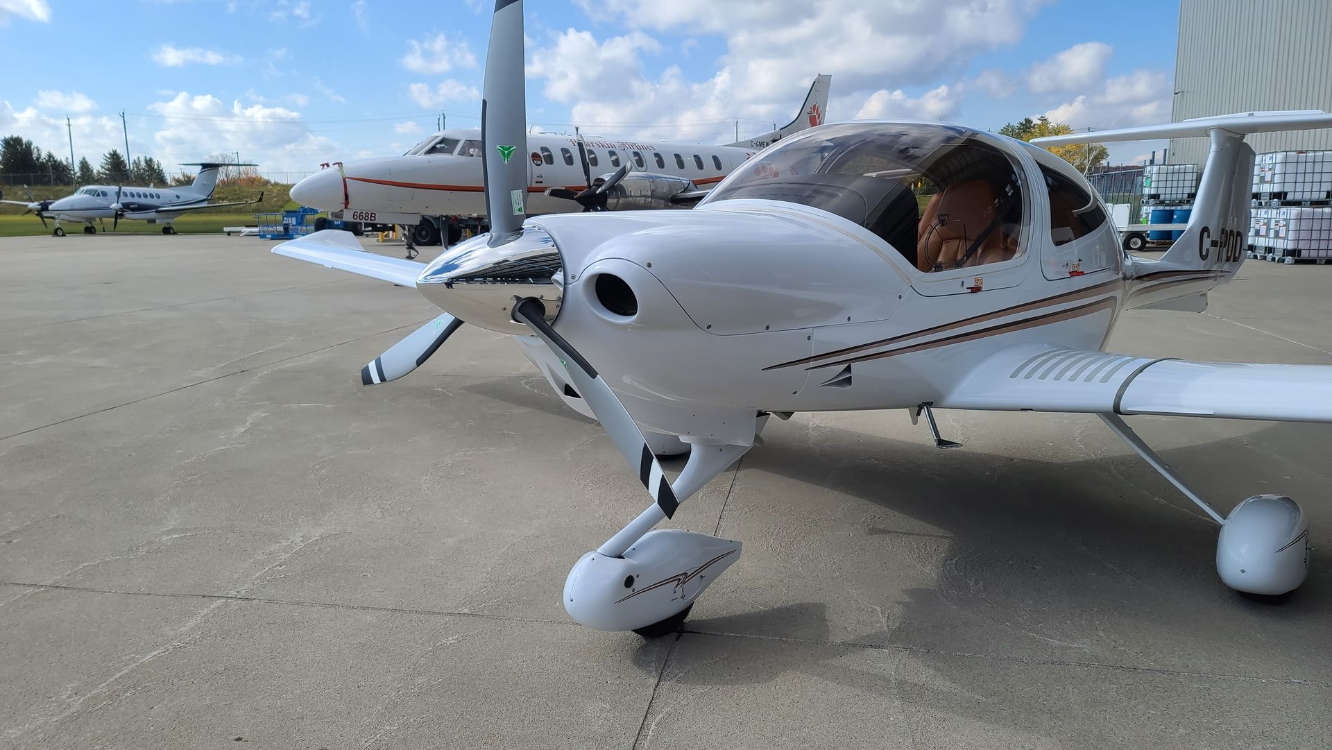White single-engine airplane with propeller on a tarmac; other aircraft in the background on a sunny day.