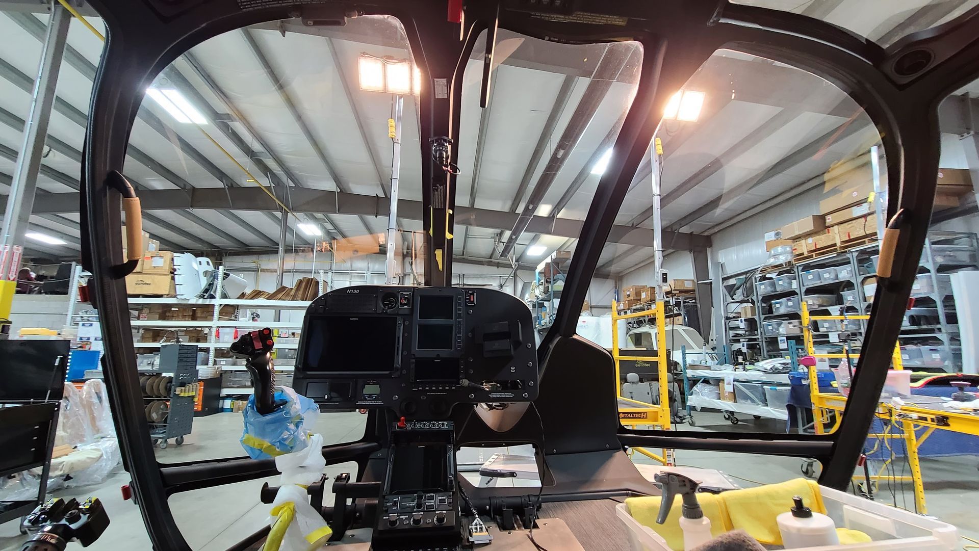 Interior cockpit view of a helicopter in a workshop, dashboard visible, with a cluttered background.
