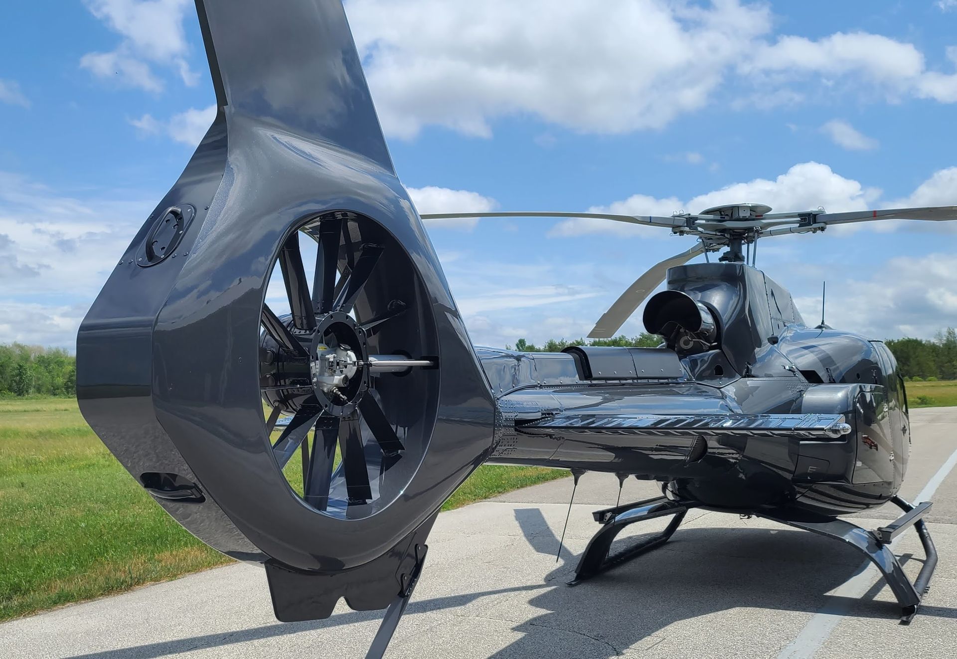 Black helicopter on a runway, tail rotor visible. Blue sky, green grass in the background.