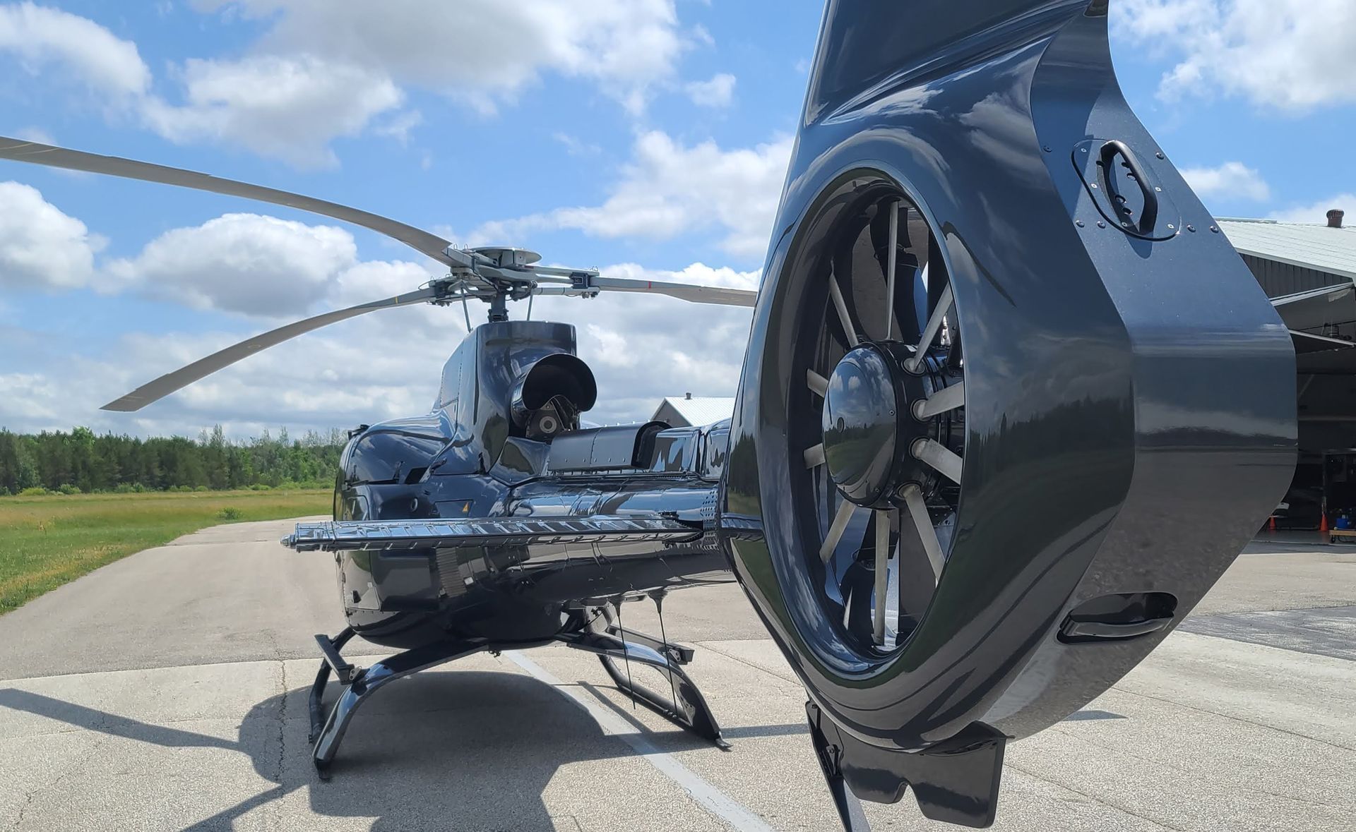 Black helicopter on a tarmac, bright sky with clouds in the background. Tail rotor and blades visible.