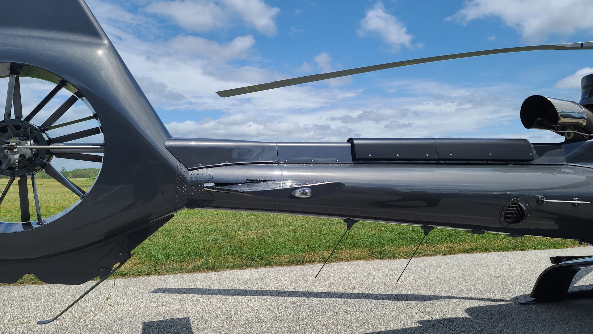 Gray helicopter tail section with rotor, on a paved surface with grass and blue sky.