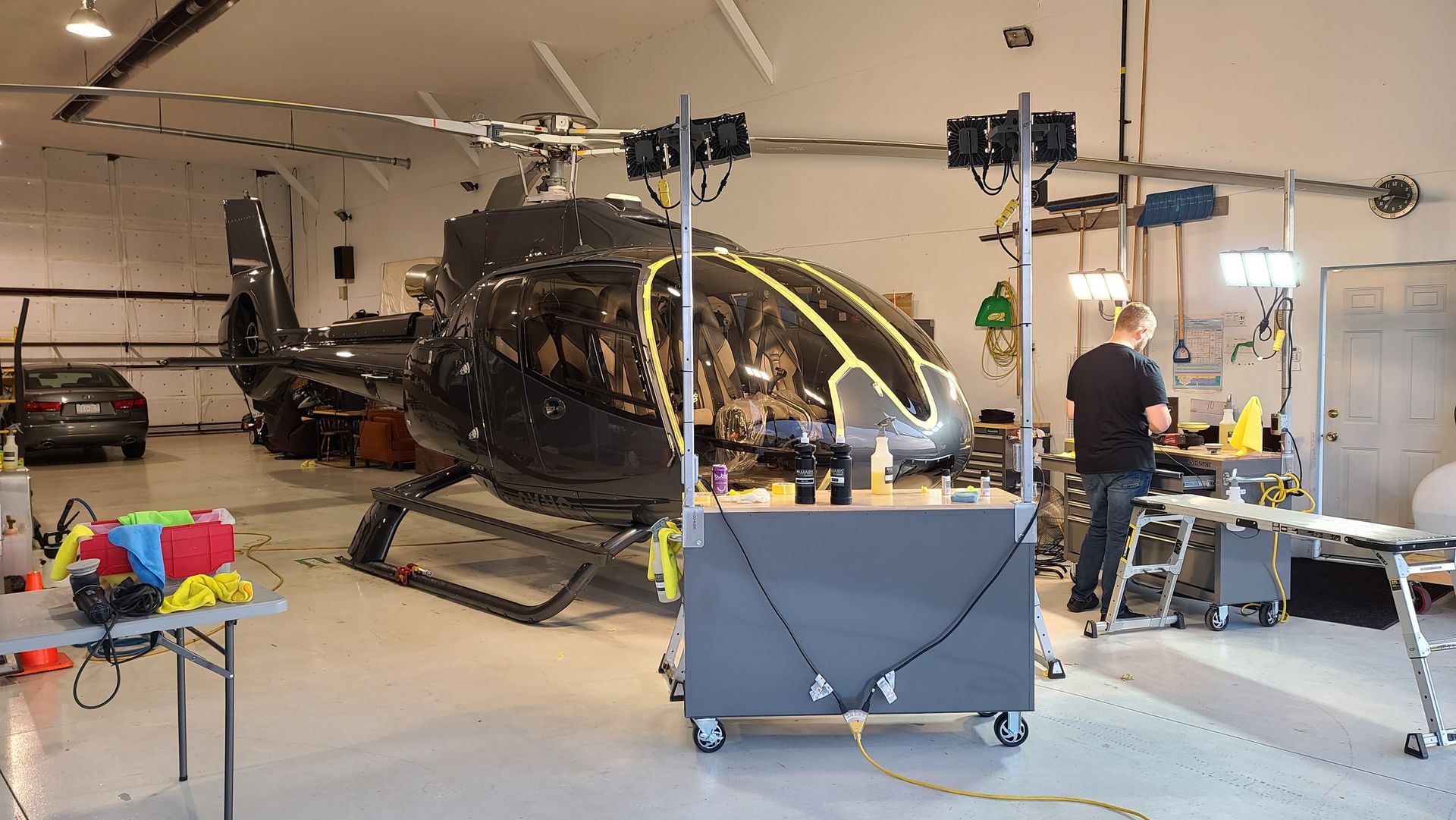 Helicopter being worked on in a hangar. Person working at a cart; lighting setup nearby.