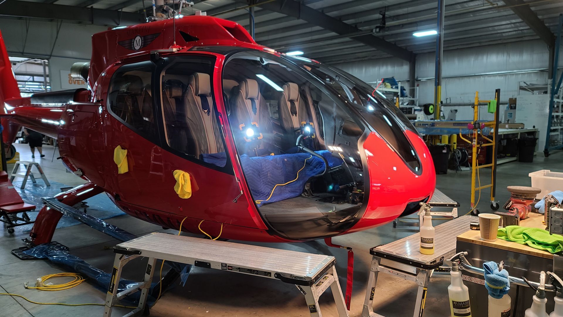 Red helicopter in a repair shop with open cockpit, surrounded by equipment and tools.