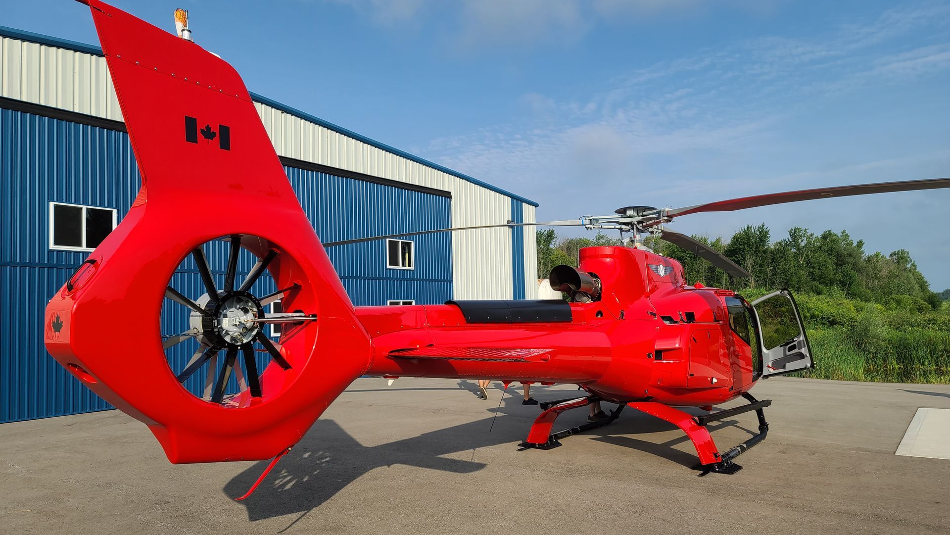 Red helicopter with Canadian flag on tail, parked near a blue building under a blue sky.