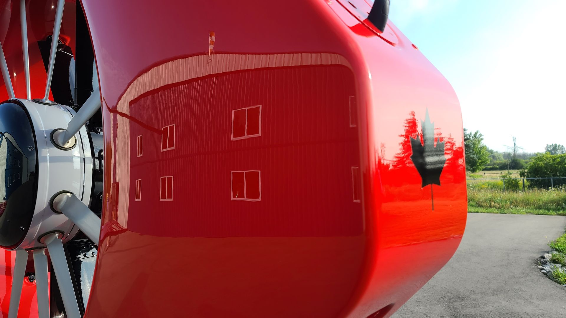 Red aircraft part reflecting a red building with windows, propeller visible, outdoor setting.