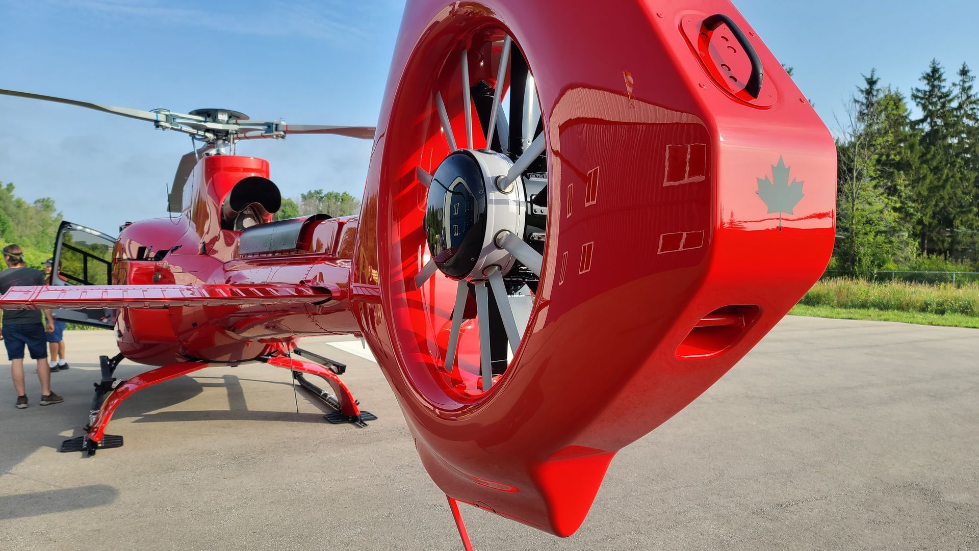 Red helicopter on the tarmac with a blurred background of people and trees under a blue sky.