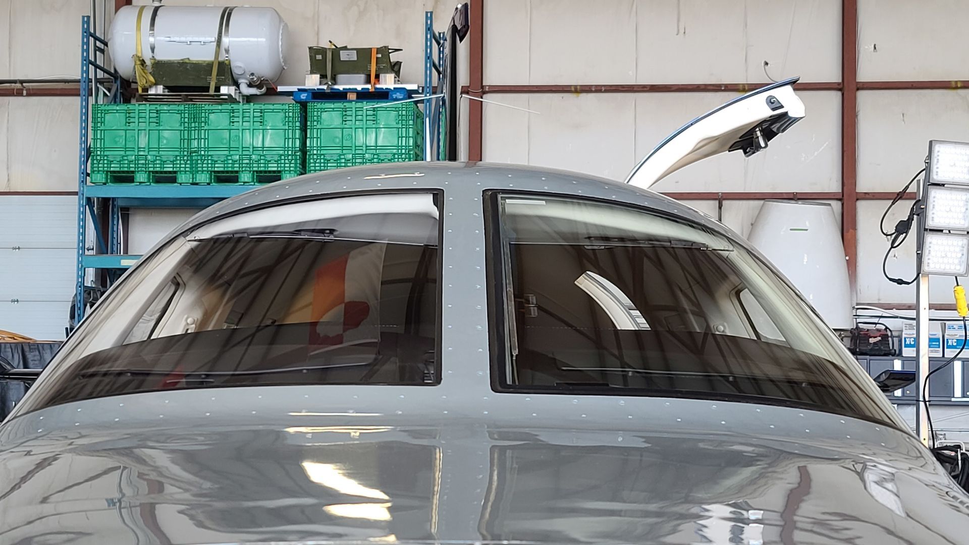 Cockpit of a gray aircraft, windshield with wipers, inside a hangar with equipment in the background.
