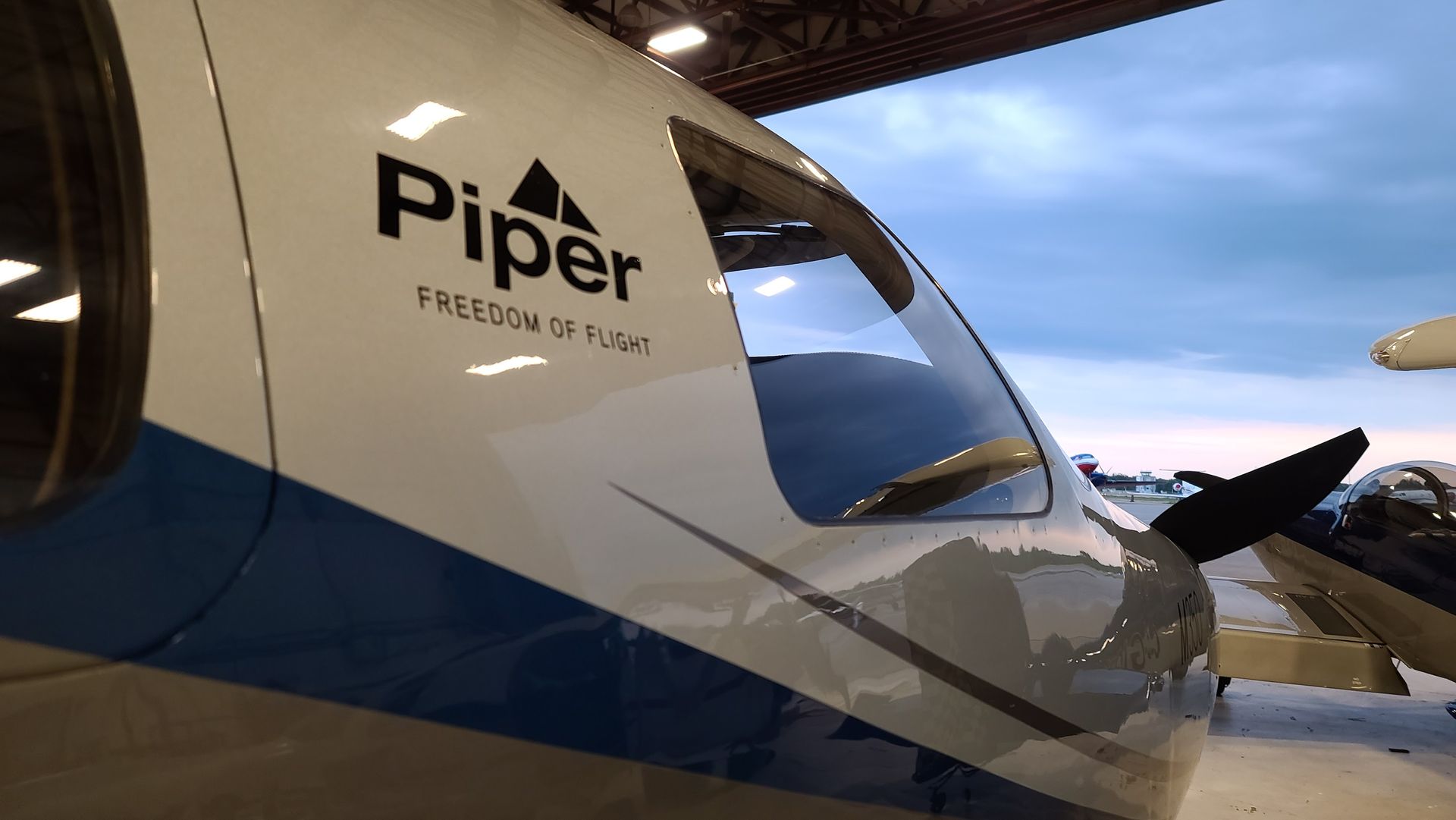 Piper aircraft cockpit with logo and windshield, blue and white paint, indoor hangar setting.