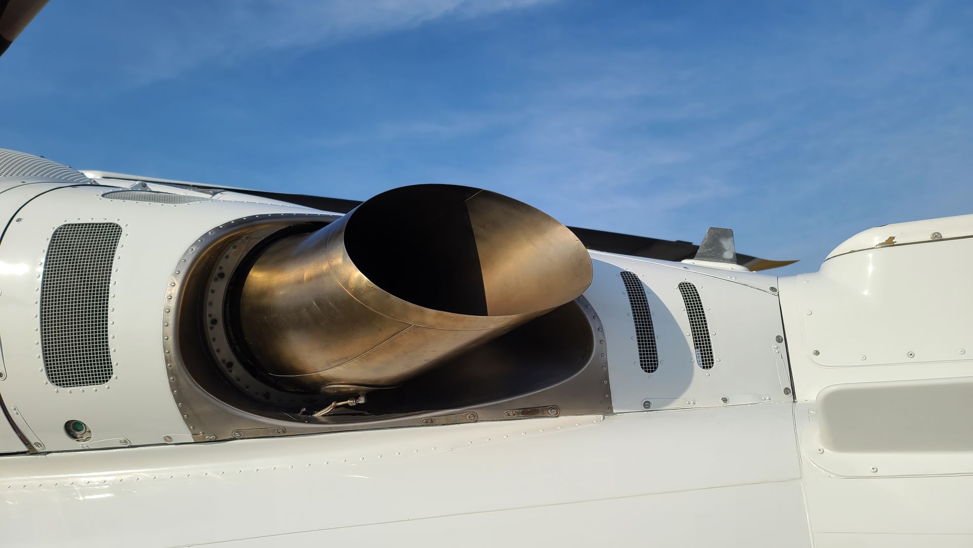A close-up view of an aircraft engine exhaust nozzle, gold-colored, set against a white fuselage and a blue sky.