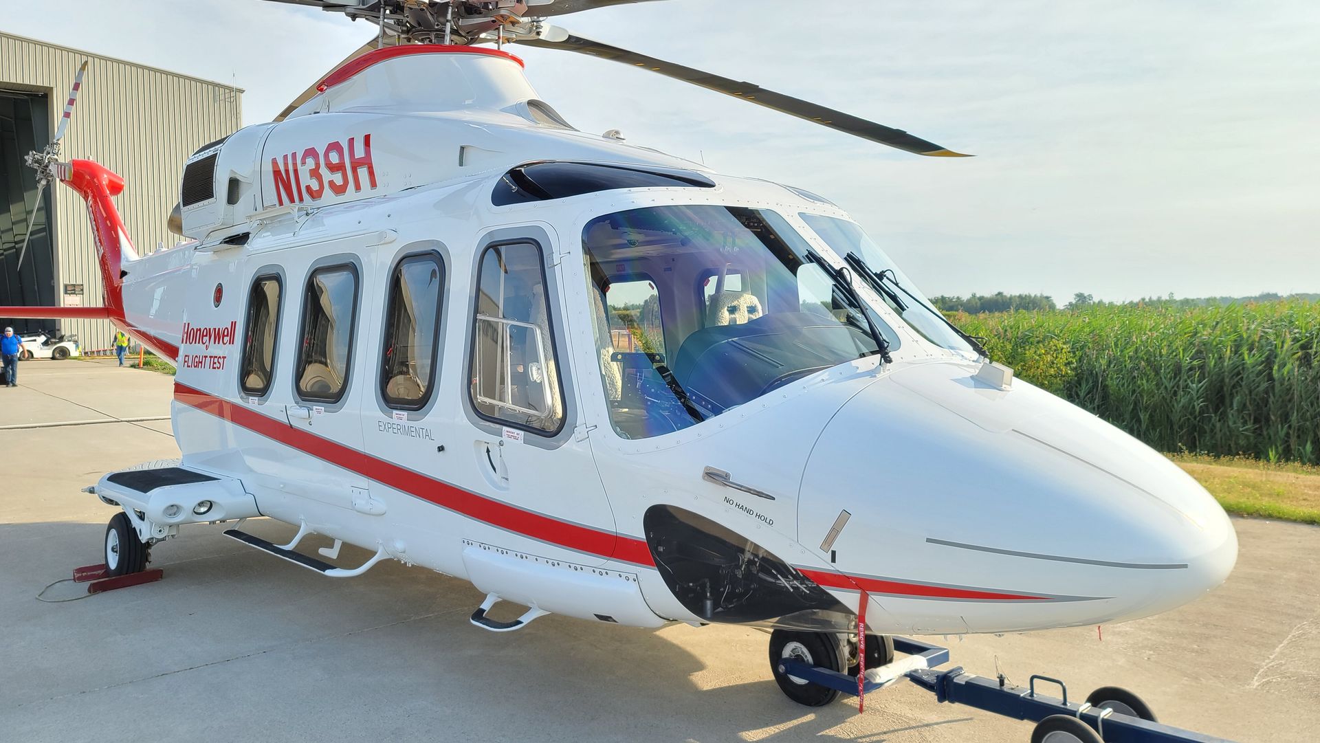 White helicopter with red accents; parked on the ground near a building and cornfield.