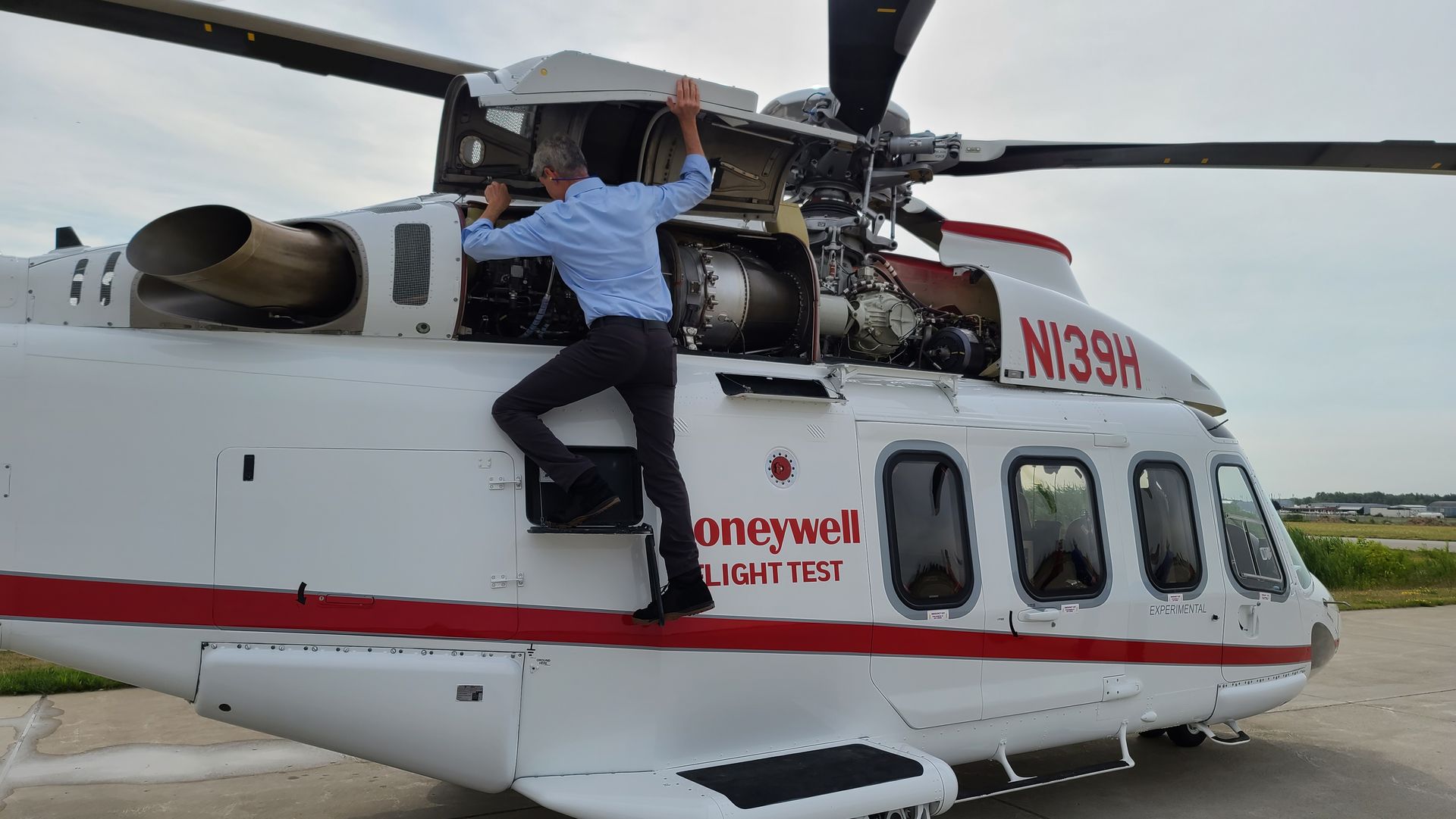 Man climbing on a white and red helicopter, next to engine, on the tarmac.