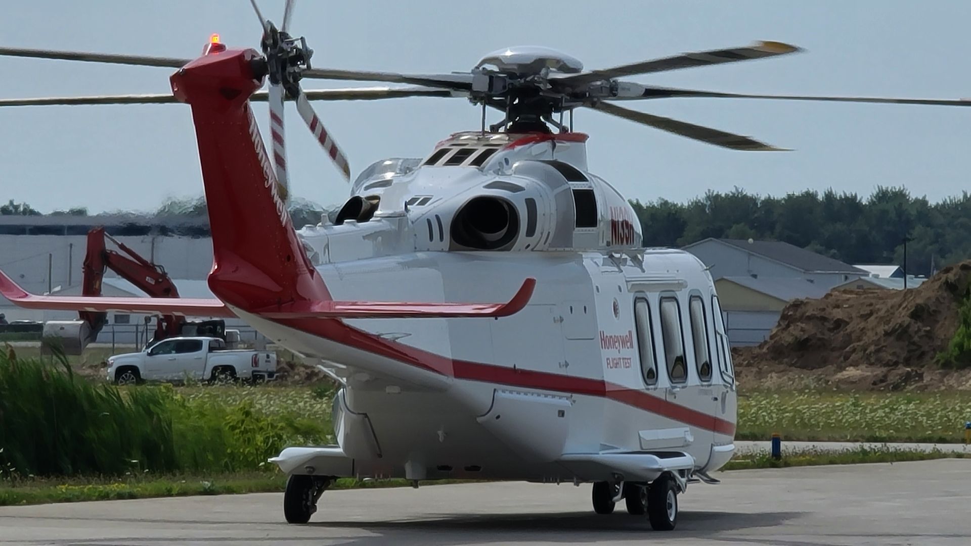 White and red helicopter on a tarmac, parked. Tail rotor visible, with a nearby truck and trees.