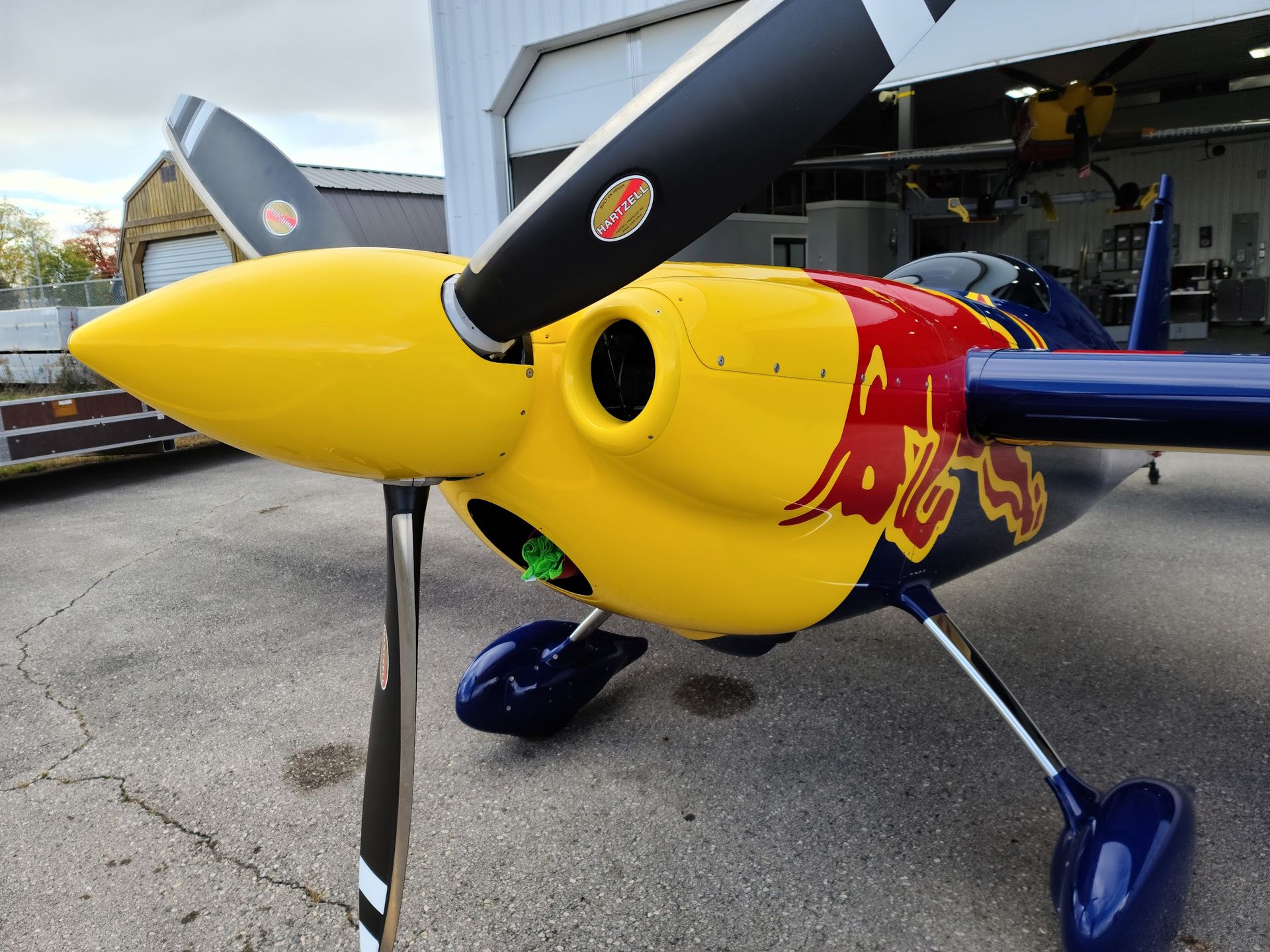 Yellow and red aerobatic plane with black propeller and red bull logo in front of hangar.