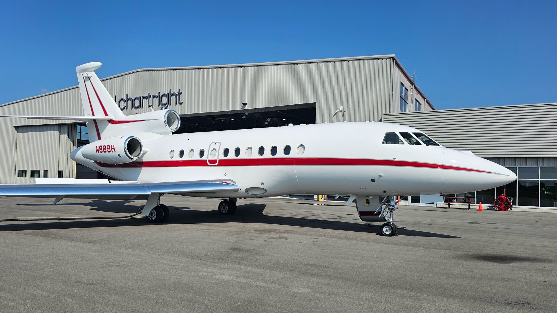 White and red private jet parked in front of a hangar on a sunny day.