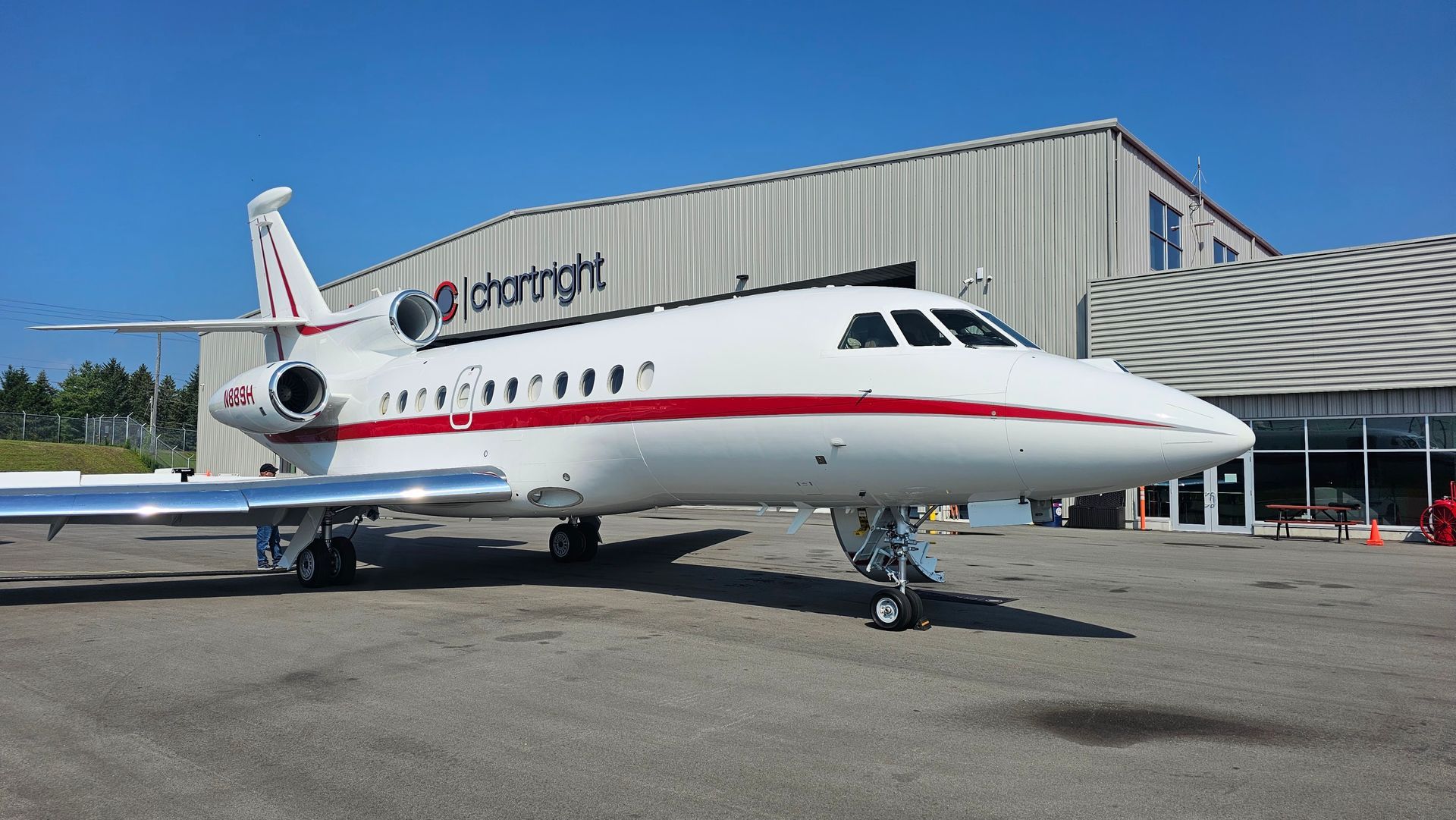 White and red private jet parked on tarmac in front of a hangar on a sunny day.