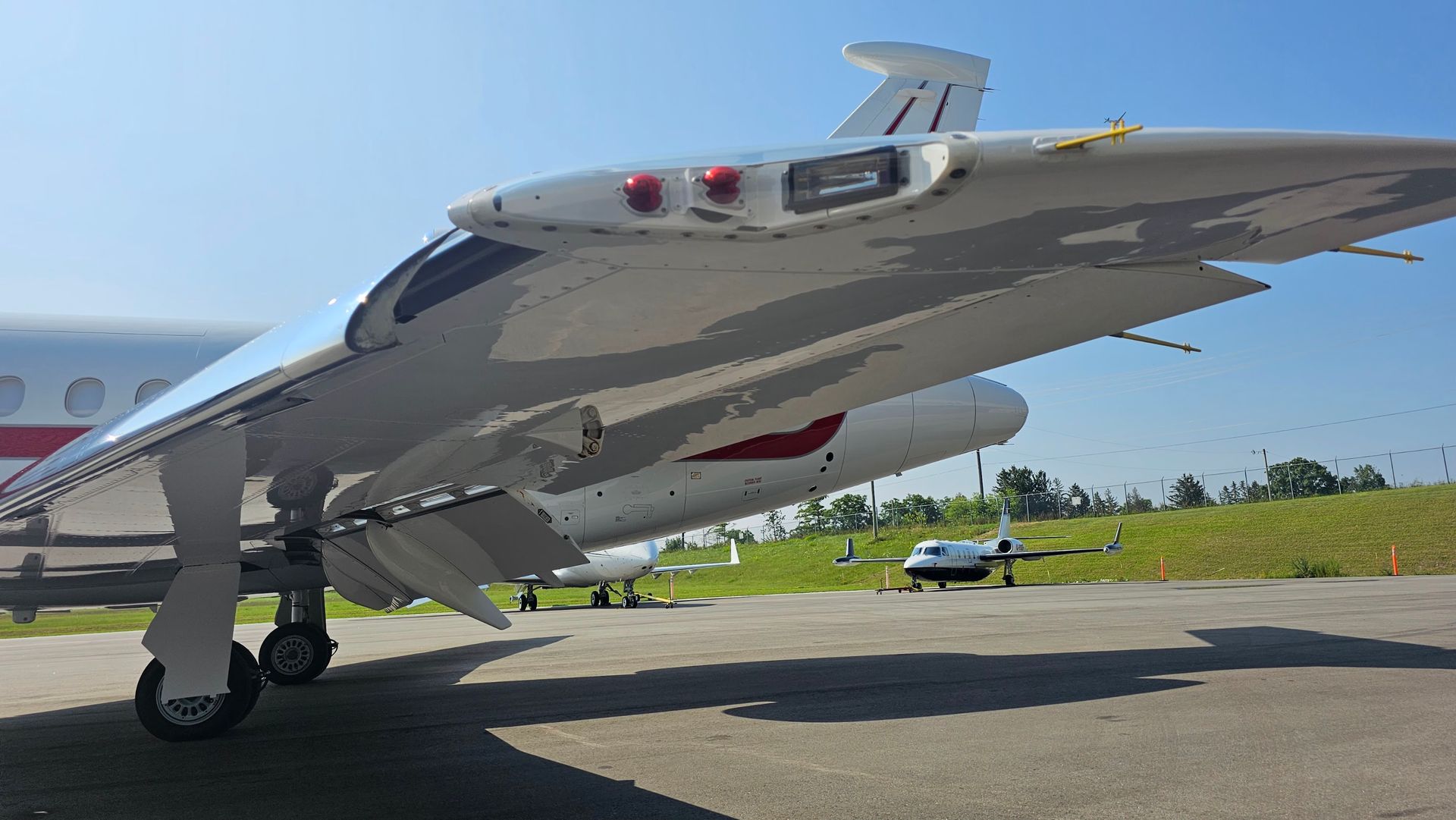 A white airplane with a red stripe sits on a tarmac, with a wing in focus and other planes visible in the background.