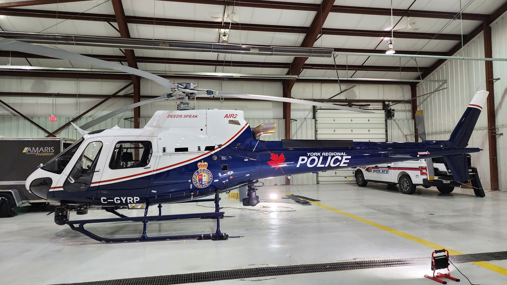 Police helicopter in a hangar, white and navy blue with the police logo; a police vehicle is parked behind it.