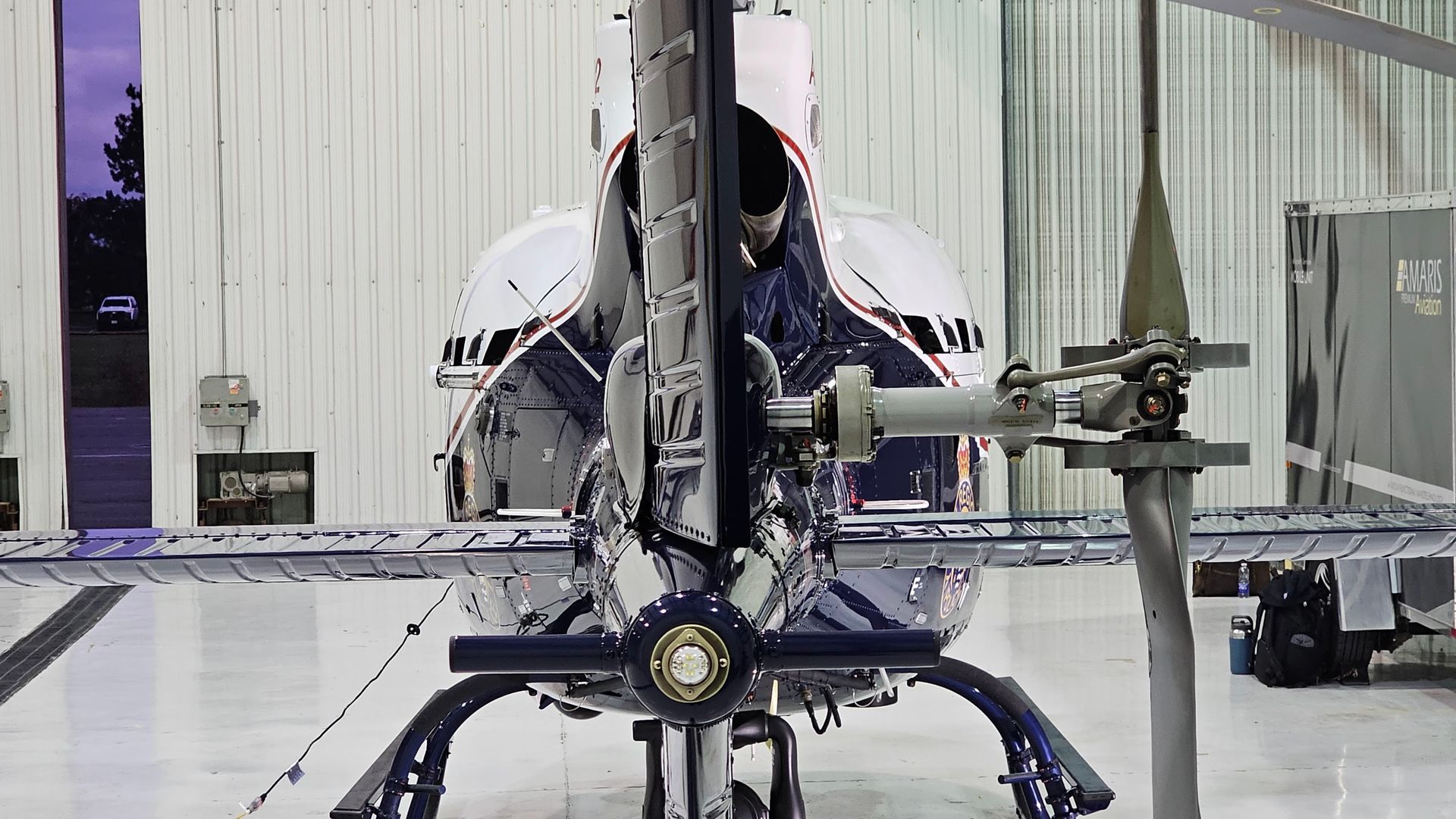 Rear view of a silver and blue helicopter inside a hangar; tail rotor visible.
