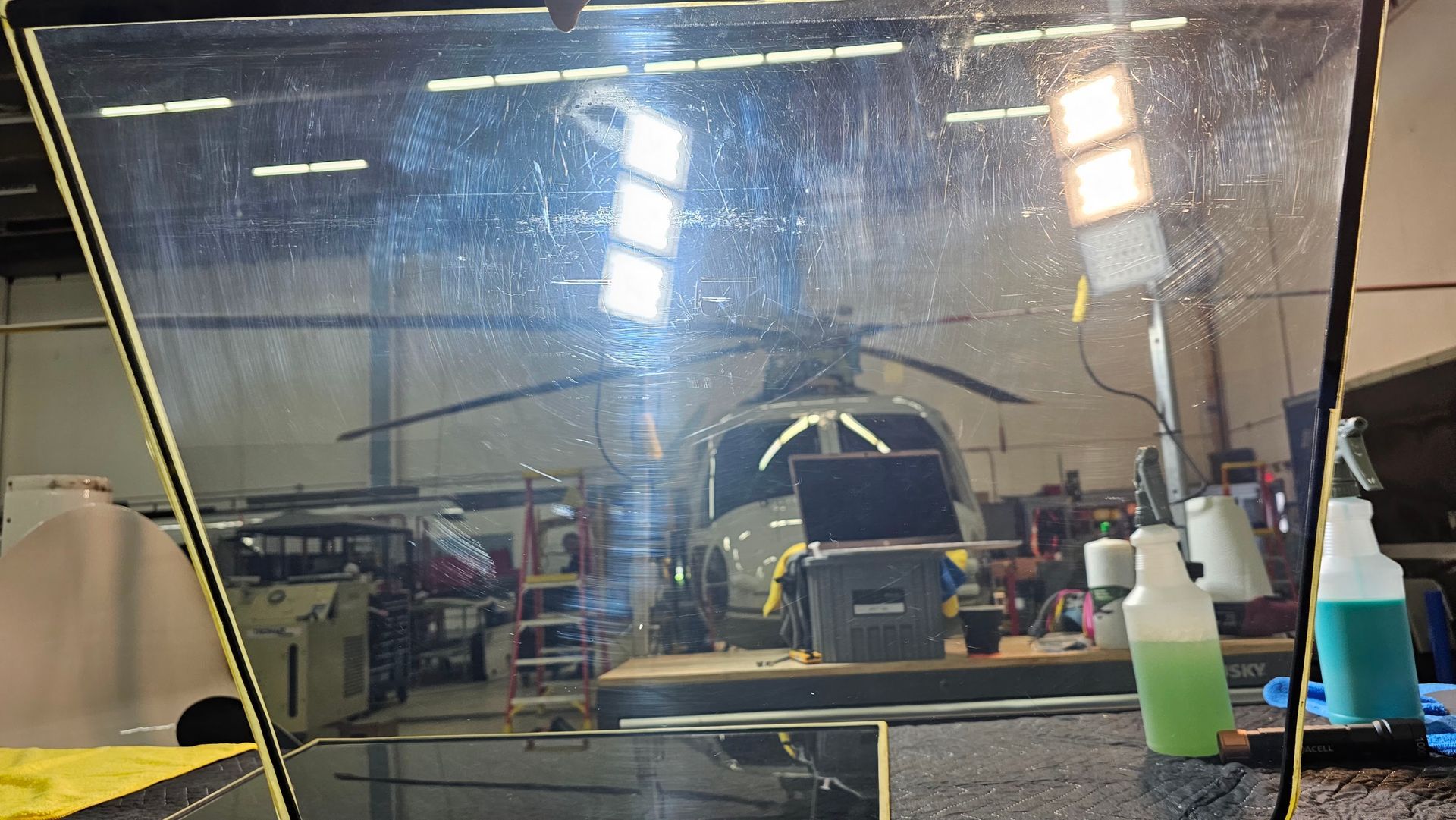 Helicopter inside a workshop, seen through a partially obscured window. Tools and bottles visible.