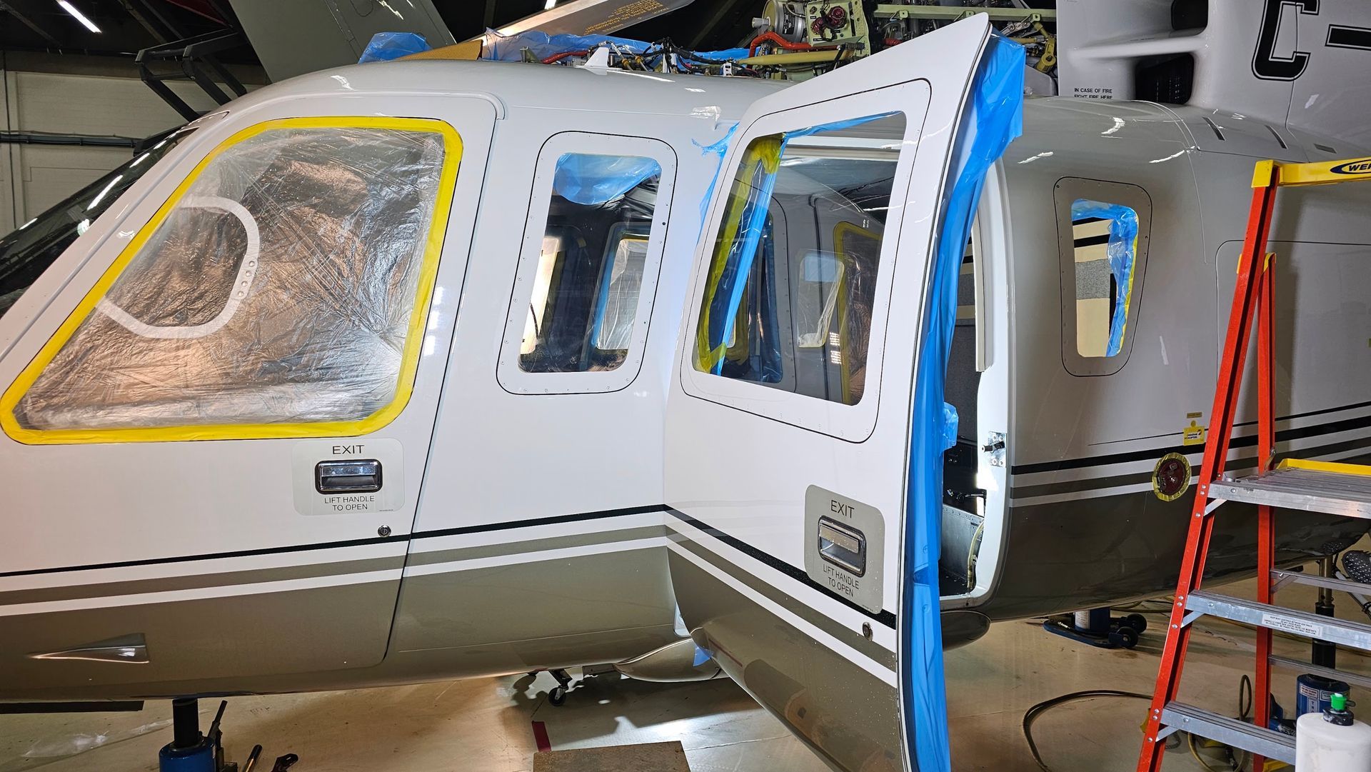 Helicopter with open door in a hangar; partially painted white with blue masking tape, a red ladder on the side.