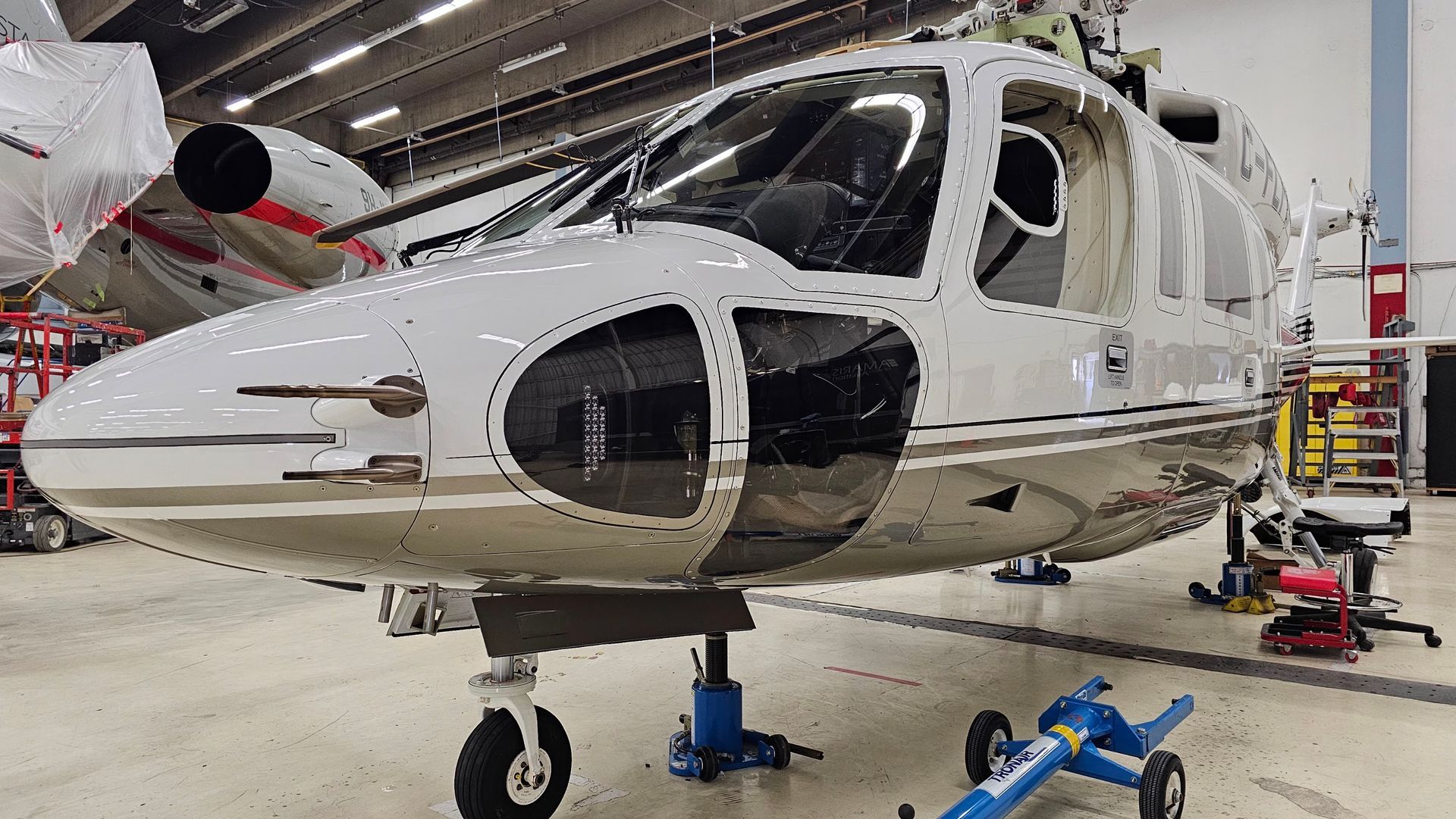 Helicopter in a hangar, white and gray with dark windows, supported by jacks on a concrete floor.