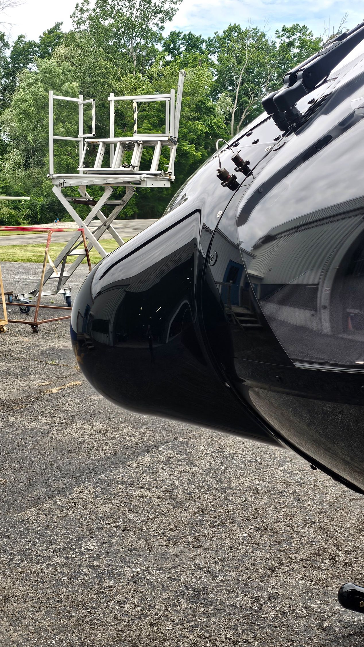 Black helicopter nose with a metal structure in the background, set on a gravel surface with trees.