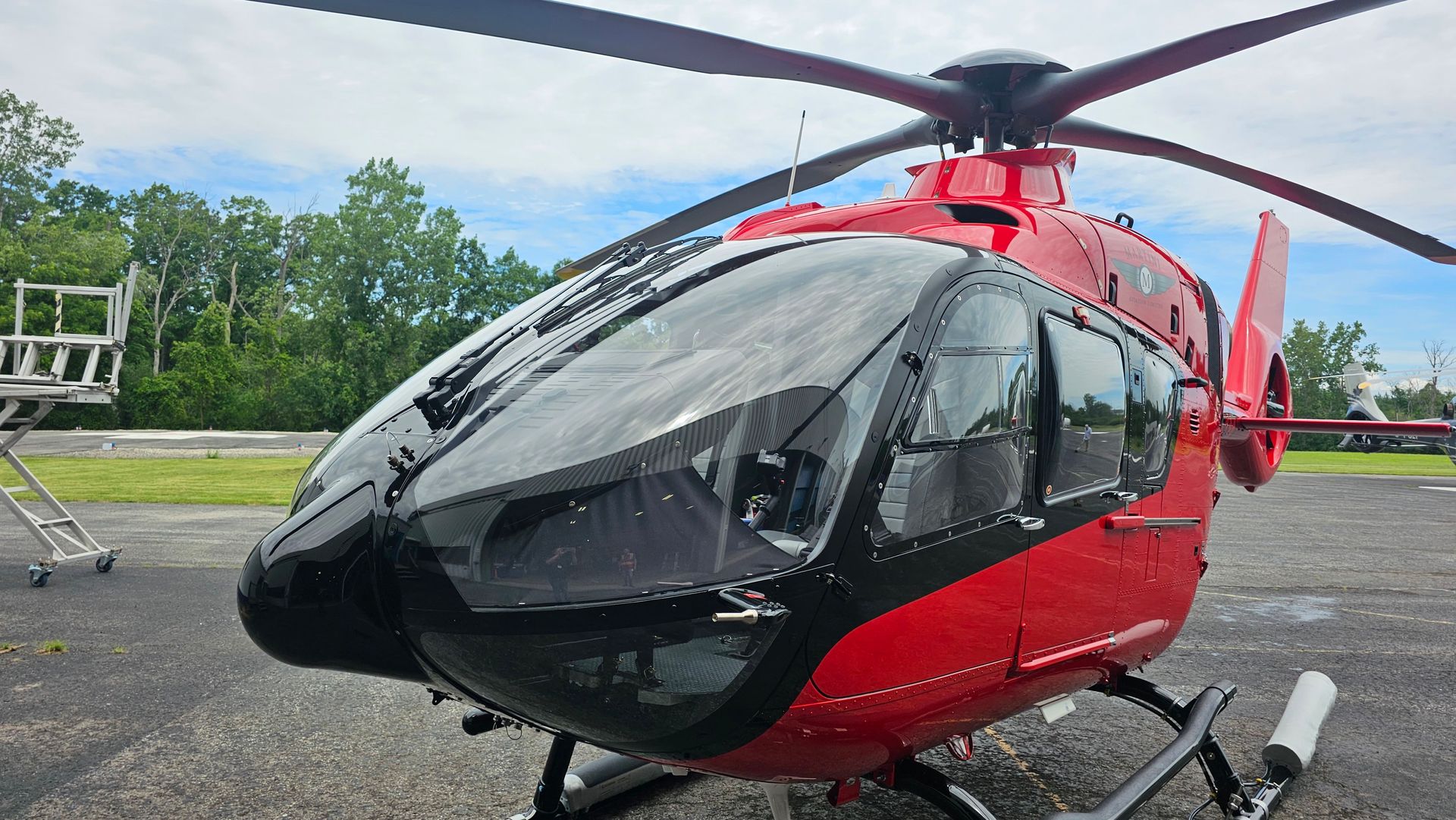 Red and black helicopter parked on tarmac with trees in the background.