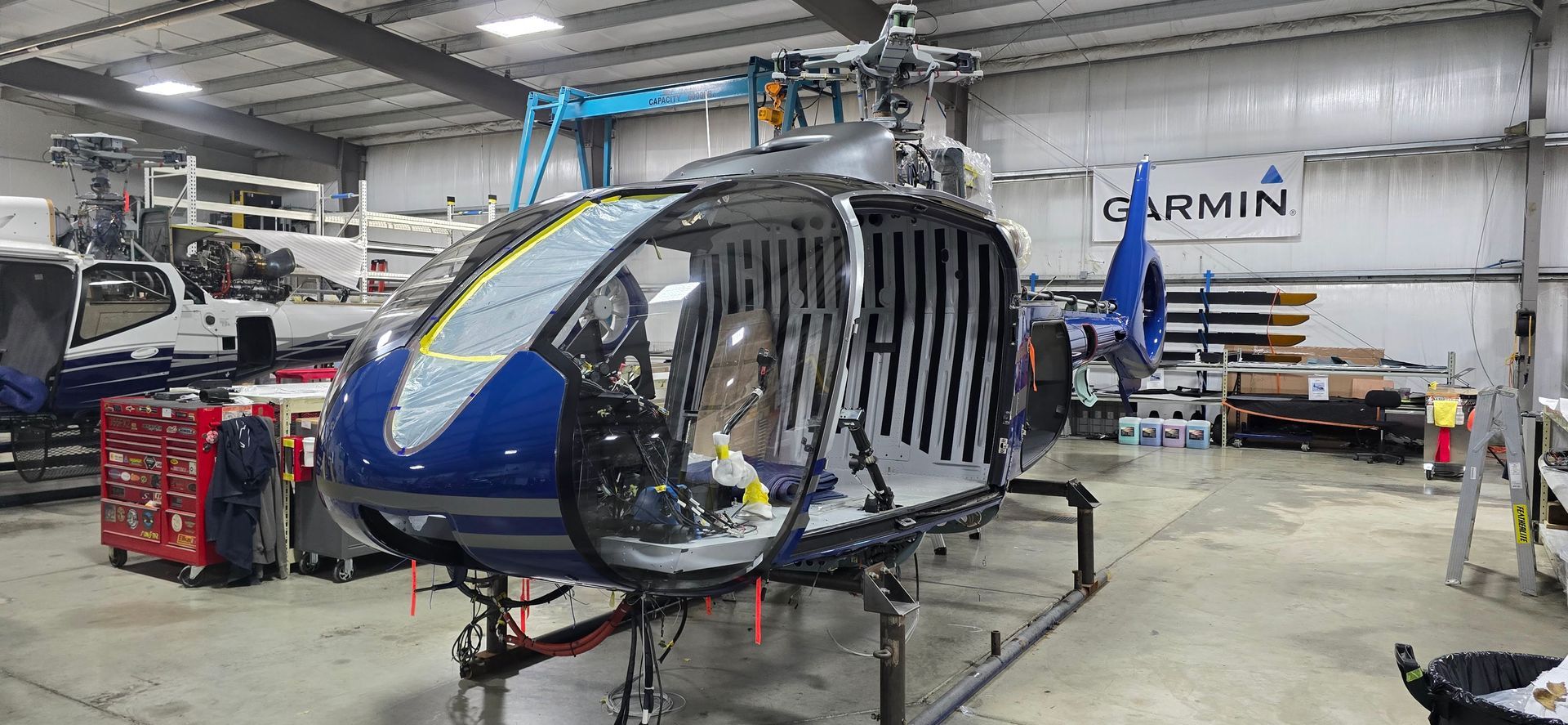 Helicopter in a workshop being worked on. Dark blue and silver. Garmin sign.
