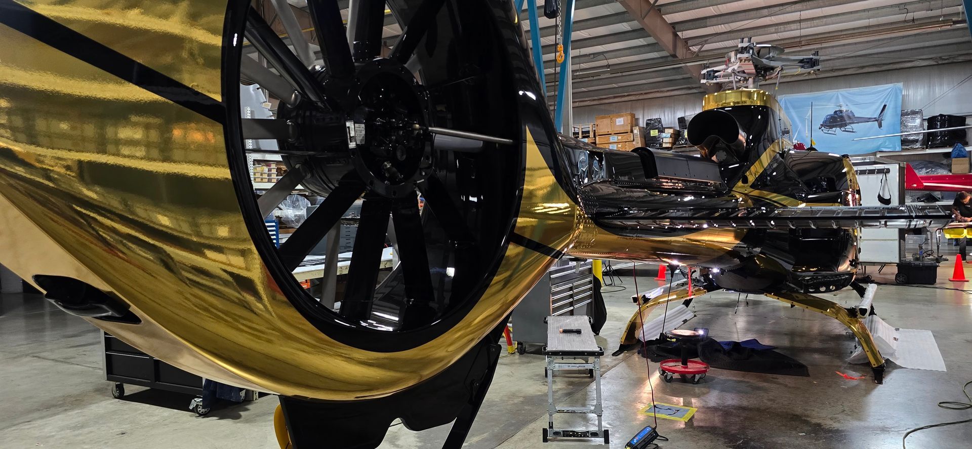 Helicopter in a hangar with gold-colored reflective panels. Black rotor and interior, workers in the background.