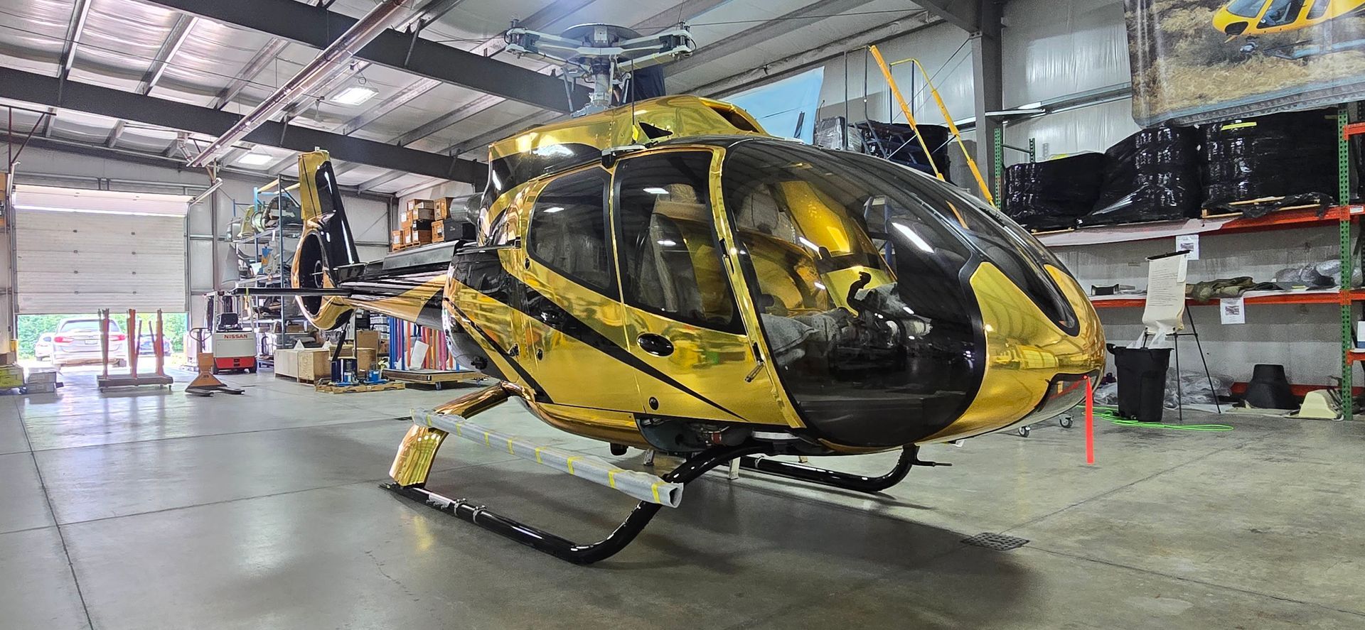 A yellow and black helicopter inside a warehouse. The hangar door is open, and shelves are visible.