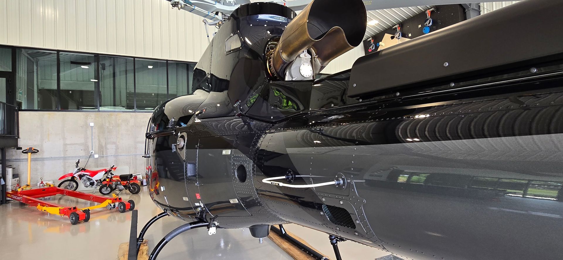 Close-up of a black helicopter in a hangar. A person works on it. Red and yellow ground equipment is visible.