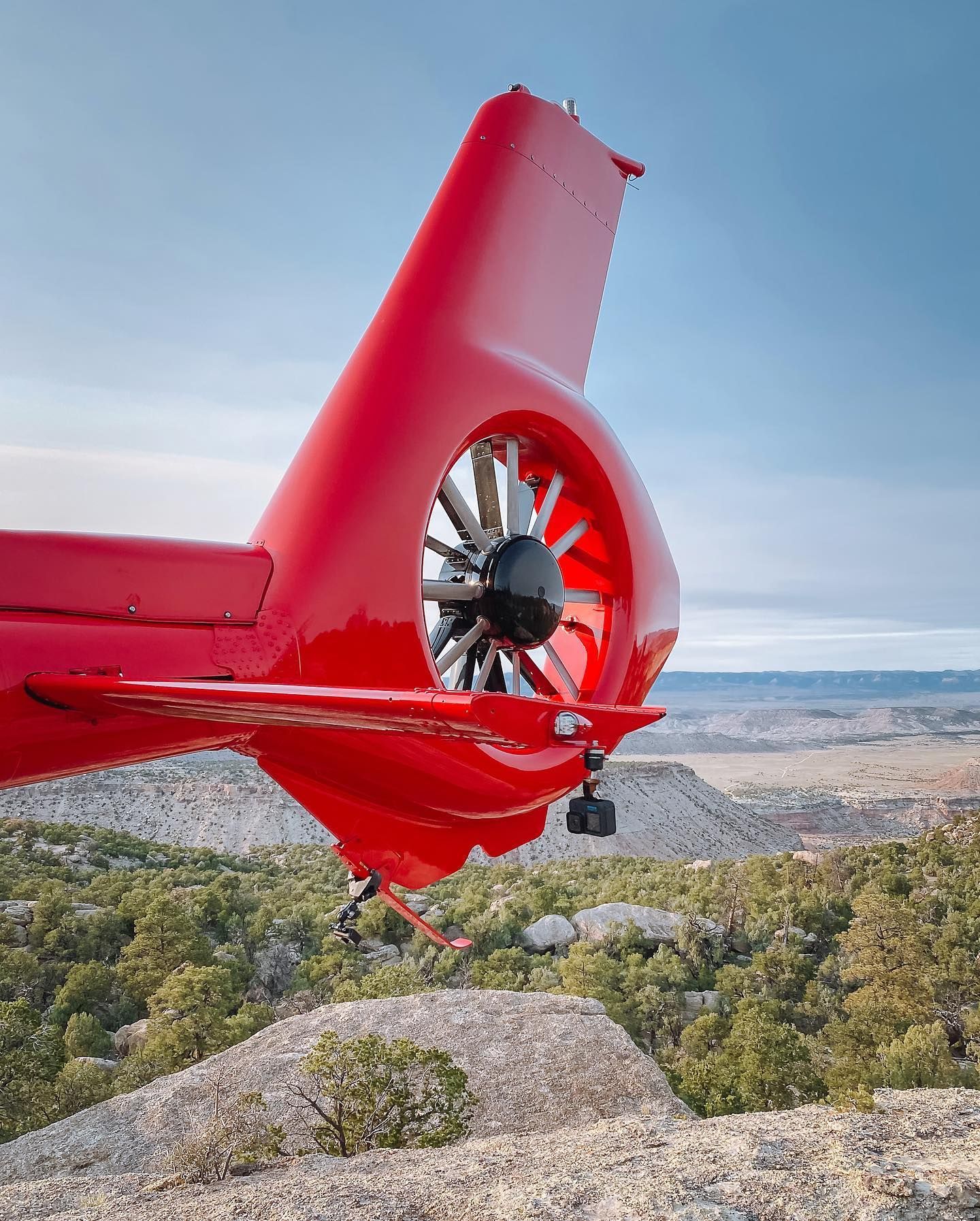 Red helicopter tail with rotor blades against a mountainous landscape and blue sky.