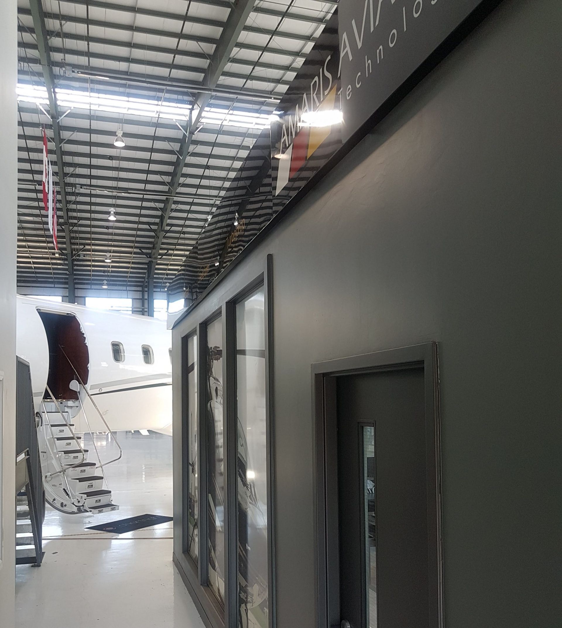 A gray wall with windows and a door inside an airplane hangar. A plane is visible.