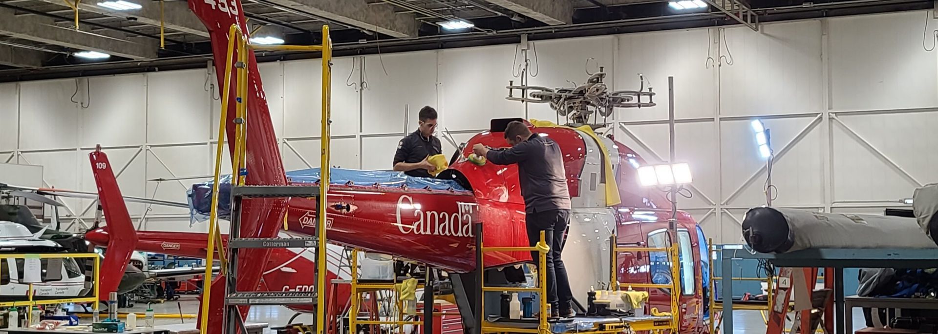 Two people working on a red helicopter inside a hangar.