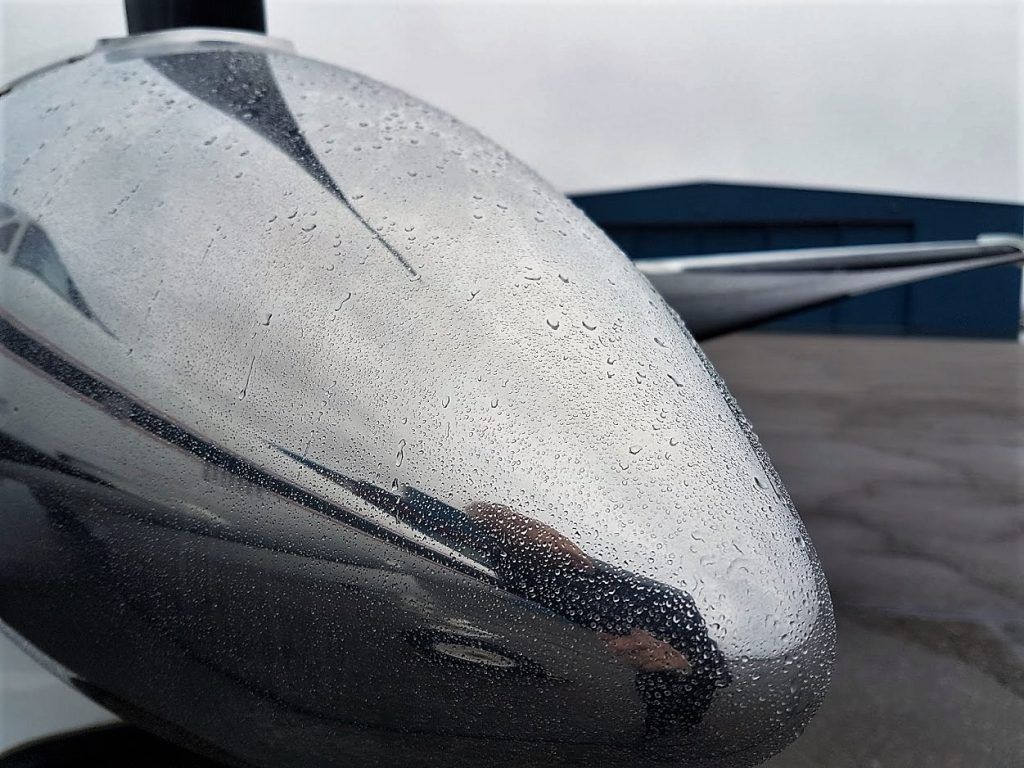 Shiny airplane nose cone covered in raindrops, reflecting sky, hangar, and wing.
