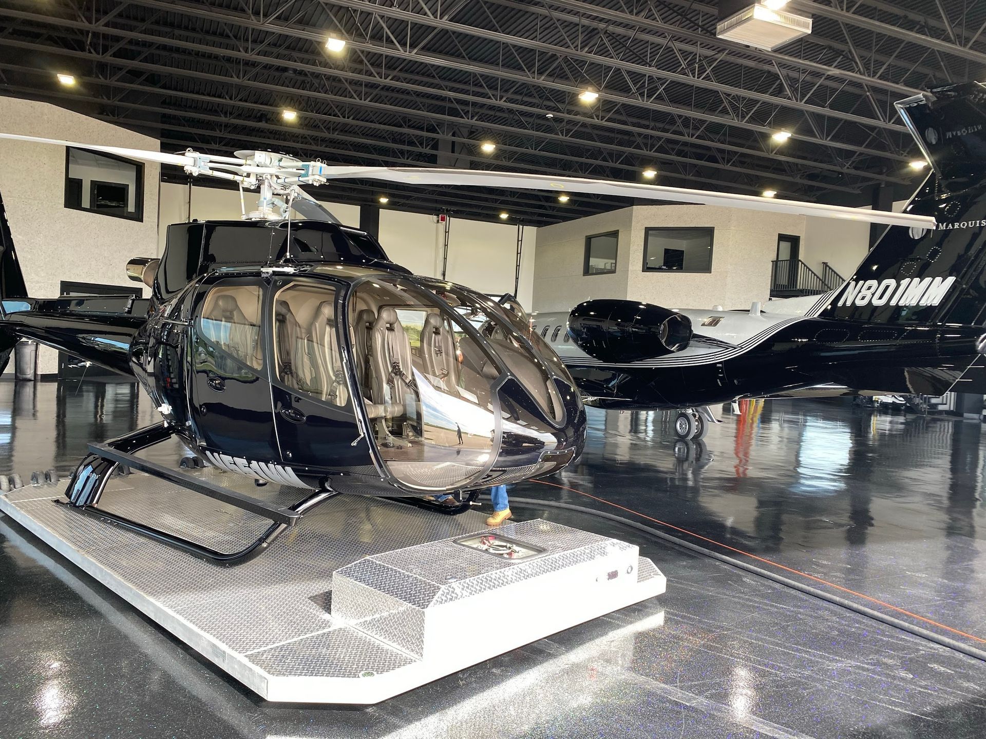 A black helicopter on a metal platform inside a hangar. Another black aircraft is in the background.