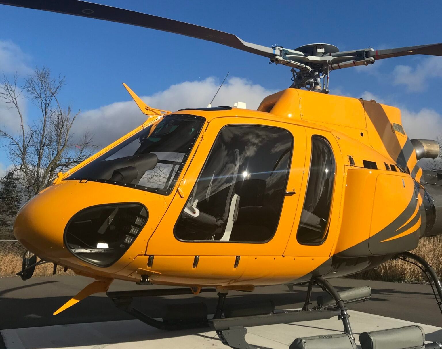 Yellow helicopter on a helipad, with black stripes and open cockpit windows against a blue sky.