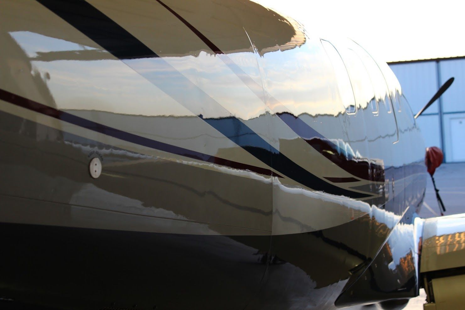 Shiny, cream-colored airplane fuselage with dark stripes; propeller visible. Reflection of a building in the surface.