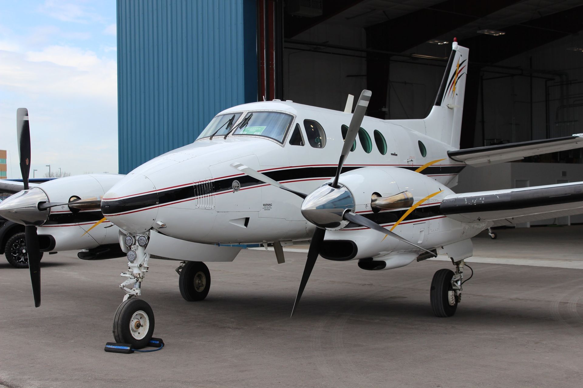 White and black twin-engine airplane parked in front of a blue hangar with its propellers and landing gear visible.