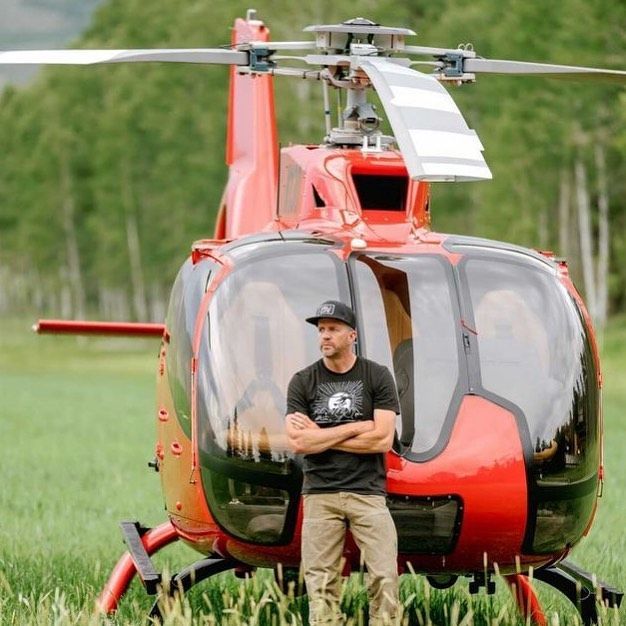 Man standing in front of a red helicopter in a grassy field.