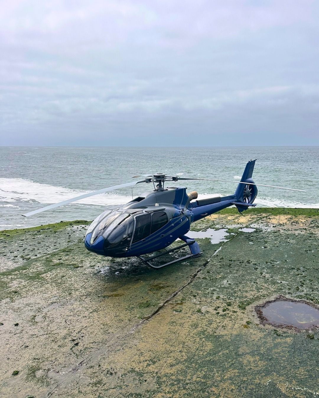 Dark blue helicopter parked near ocean shoreline under cloudy skies.