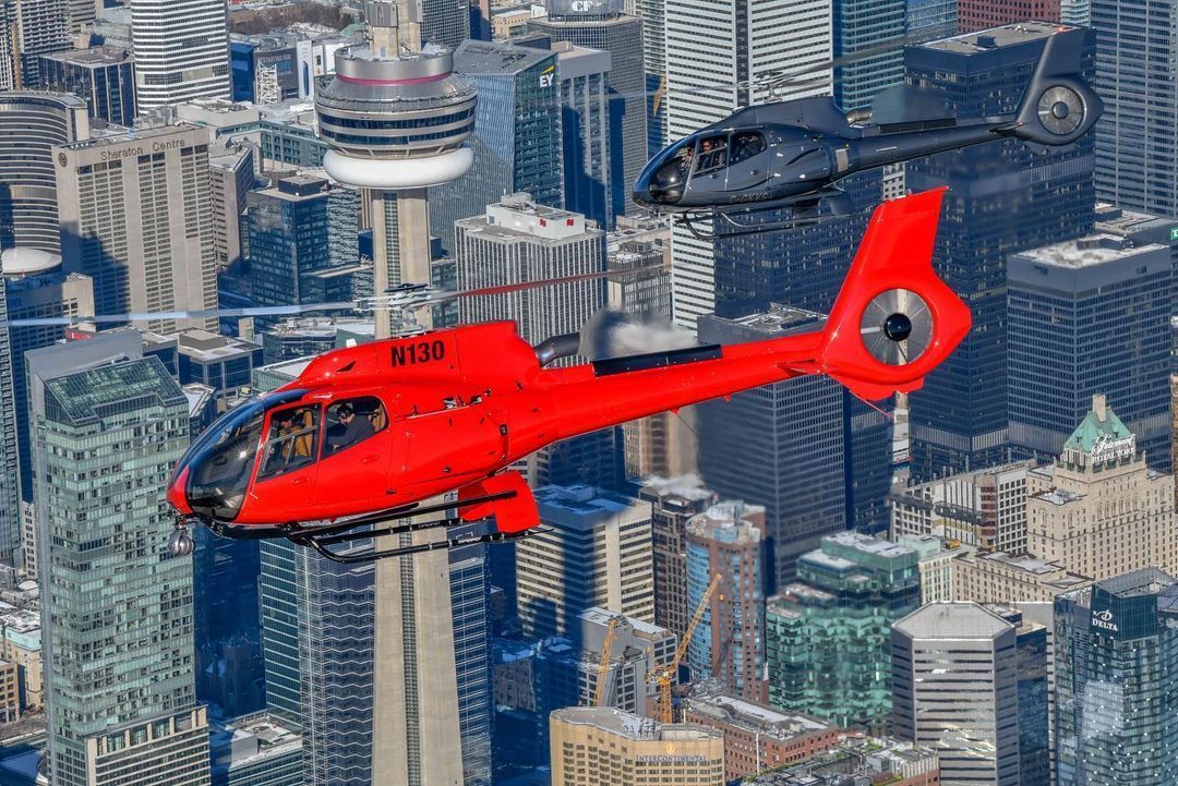 Two helicopters flying over a cityscape with the CN Tower. The lead helicopter is red; the other is black.