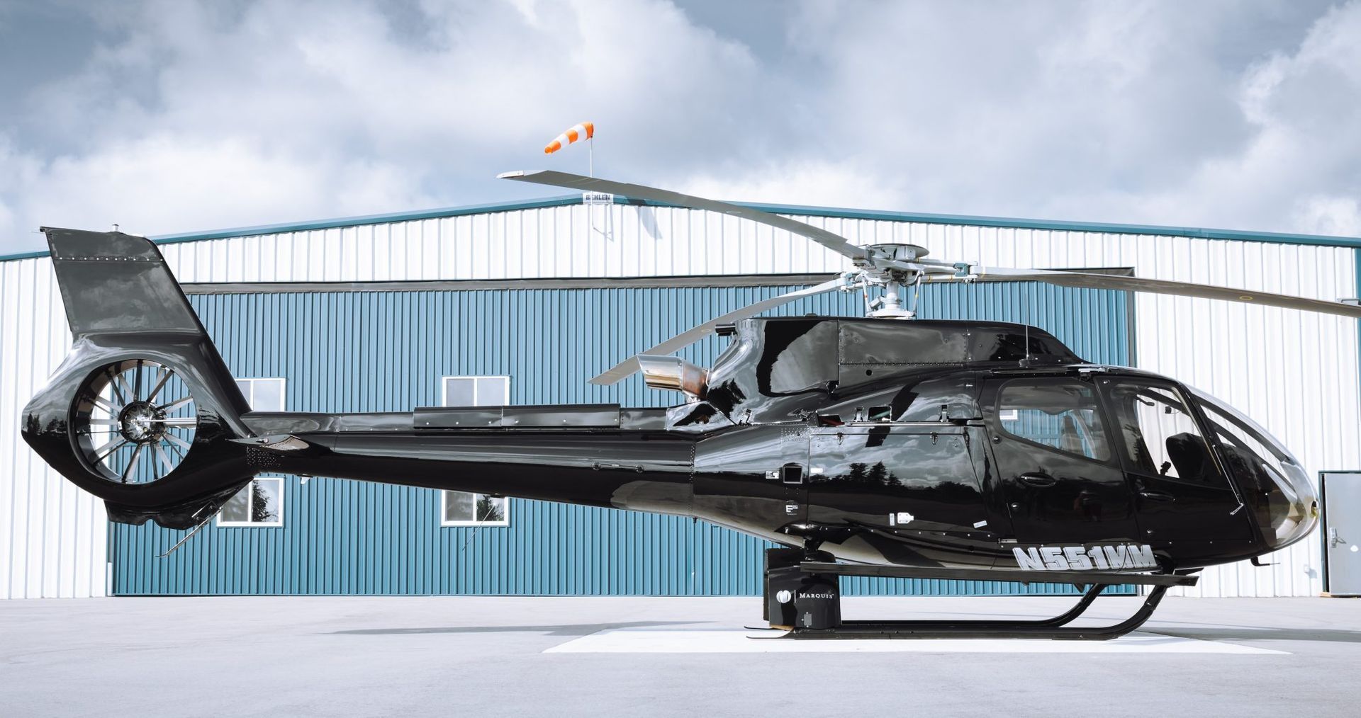 Black helicopter parked on a concrete pad in front of a blue and white building, under an overcast sky.