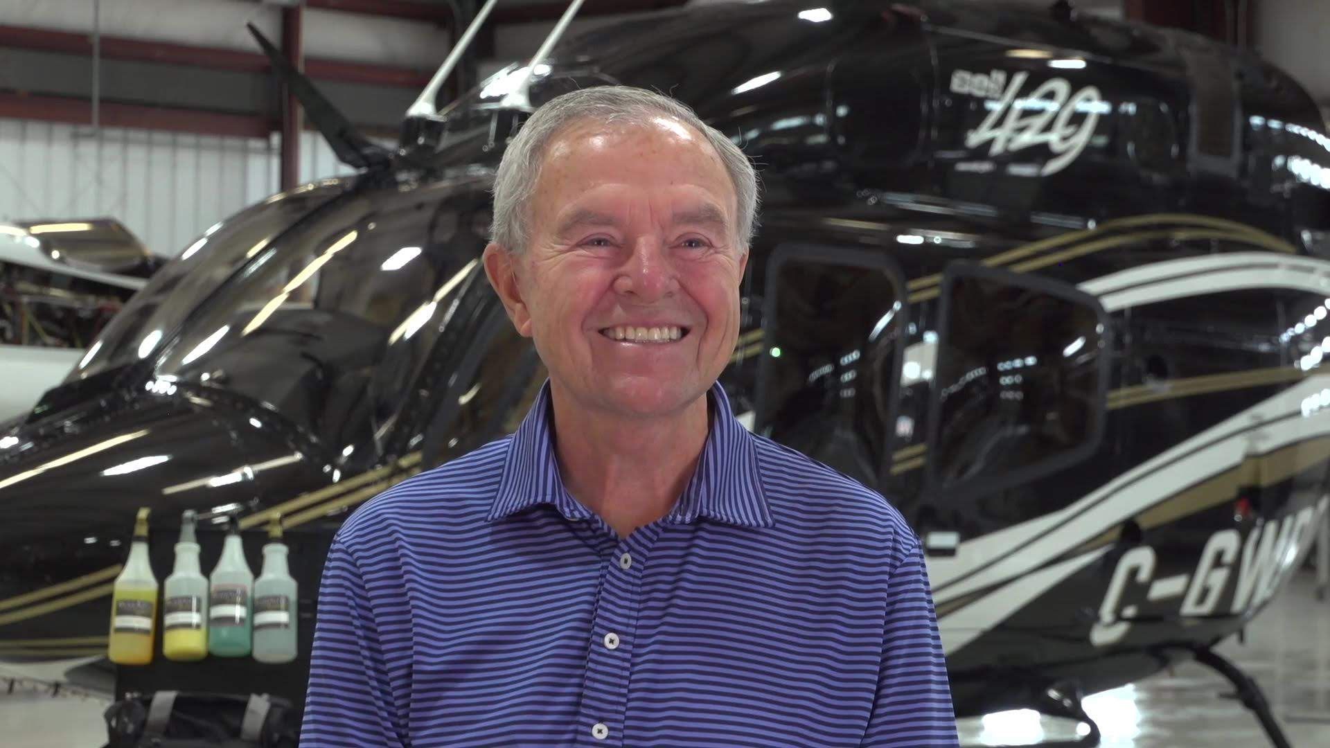 Man smiles in front of a black helicopter with gold accents; indoor hangar setting.