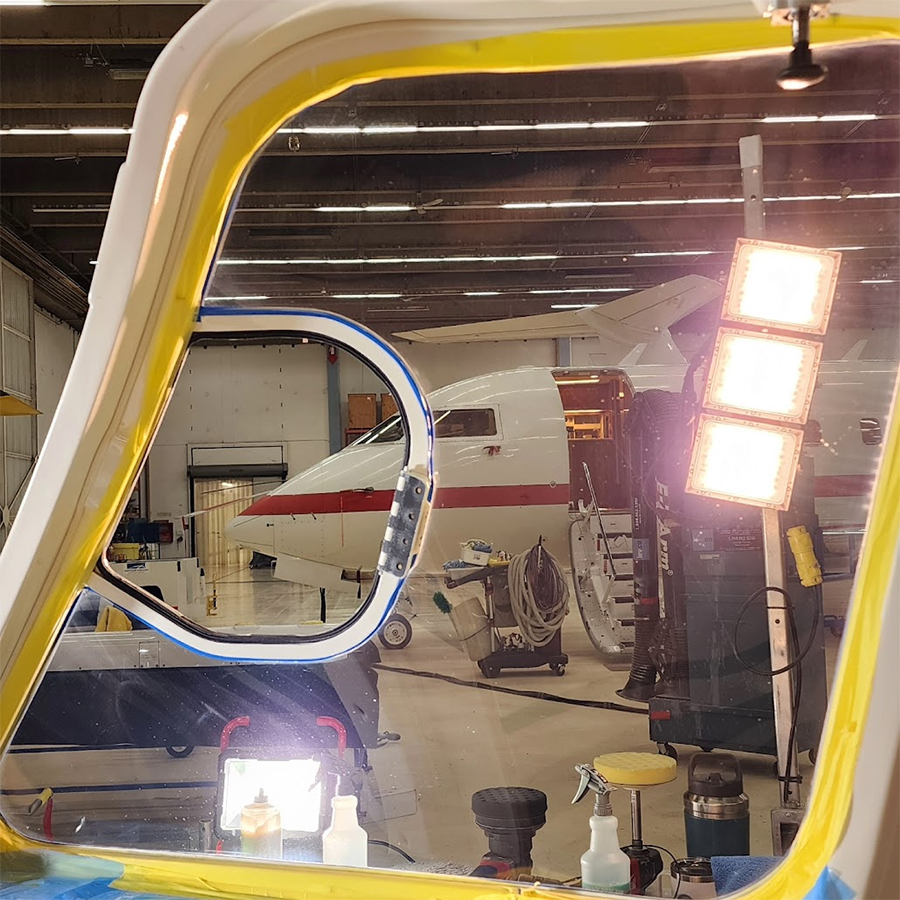 Inside a hangar, a plane is seen through an open window frame. It has a white body with red accents.