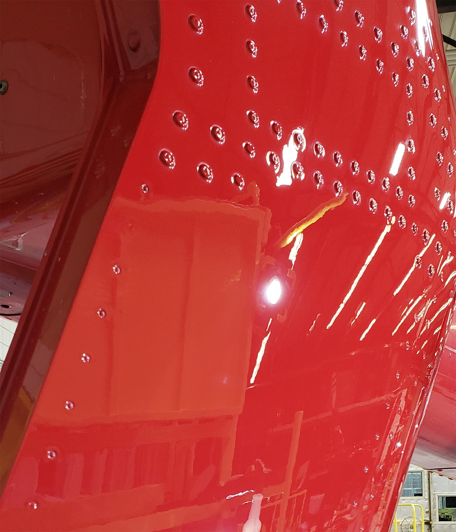 Shiny red aircraft panel with rows of rivets, reflecting light and surroundings.